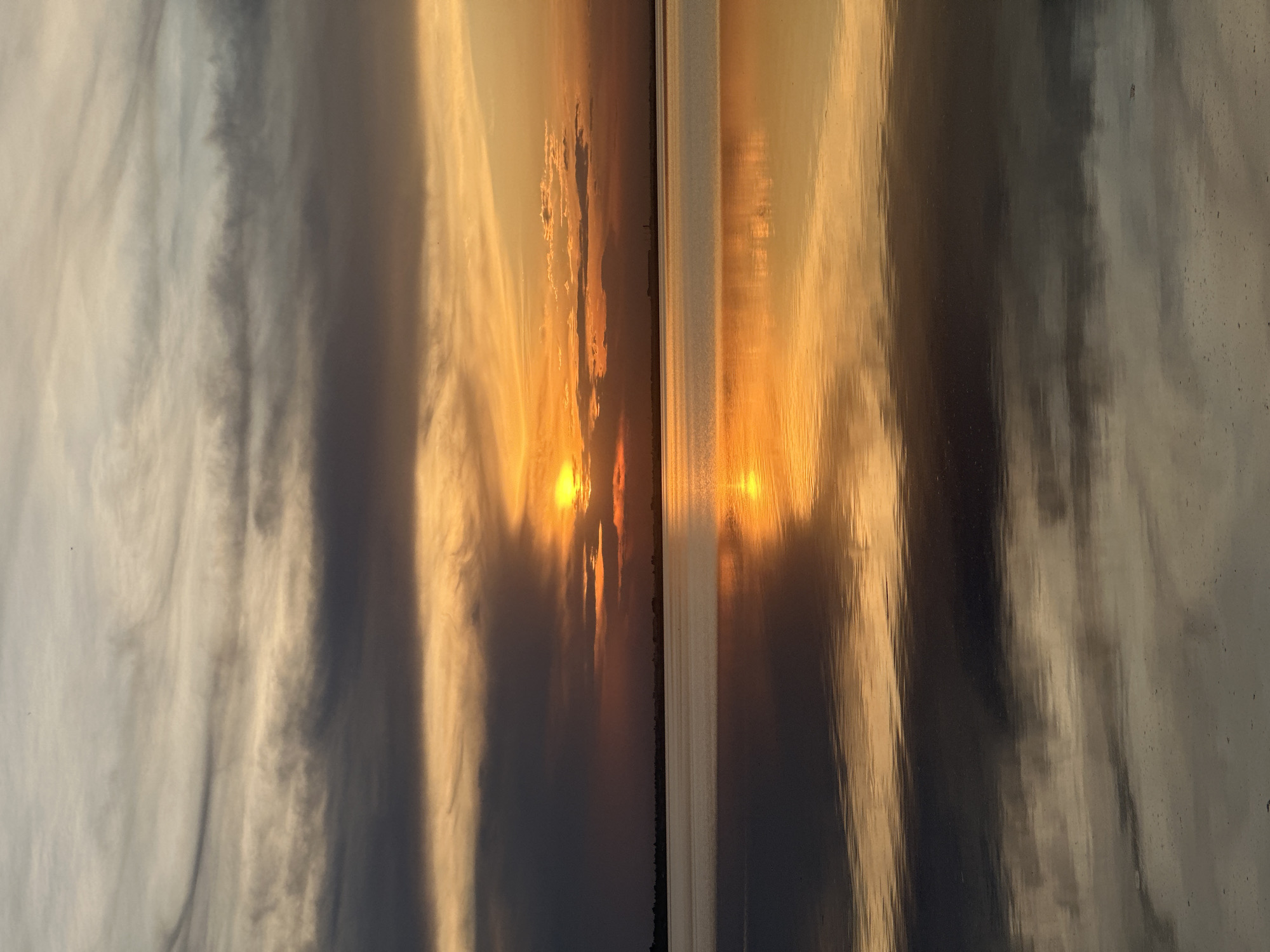 The image shows a sunset reflecting on a body of water. The sky is filled with clouds, and the setting sun casts a warm orange glow. There appears to be a shoreline or landmass in the distance. The reflection on the water mirrors the sky and sun. The location is Kokshetau, Kazakhstan. The photograph is turned on its side so the water looks like land.
