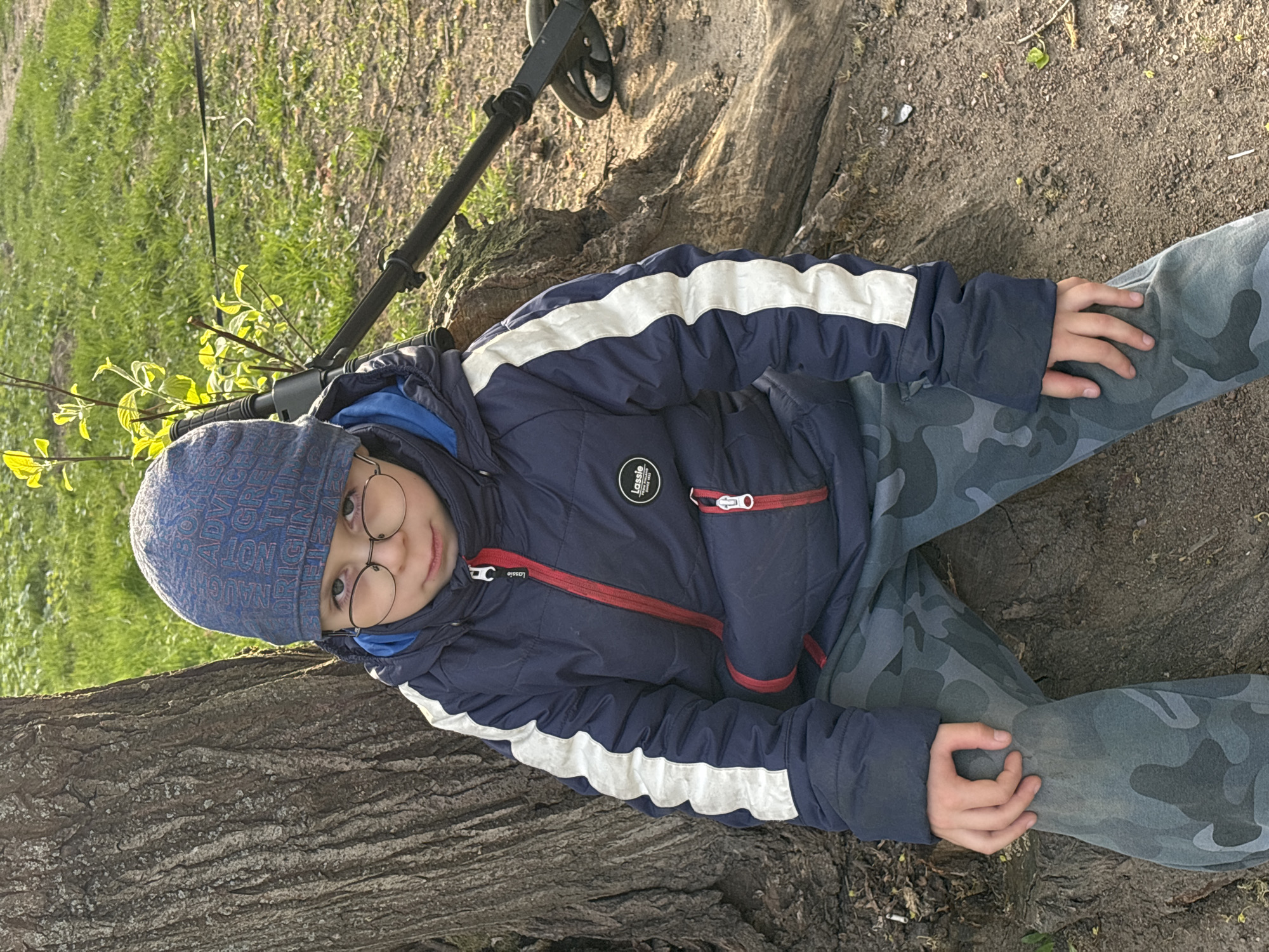 A young boy with round glasses, wearing a blue winter jacket with white stripes and camouflage pants, sits on a large tree trunk. He is wearing a blue beanie with text on it. Behind him is a metal detector resting on the trunk and ground. The ground is dirt with patches of grass. The photo appears to be taken outdoors in a park-like setting. It is not possible to determine the location specifically in Saint Petersburg, Russia, without further information.