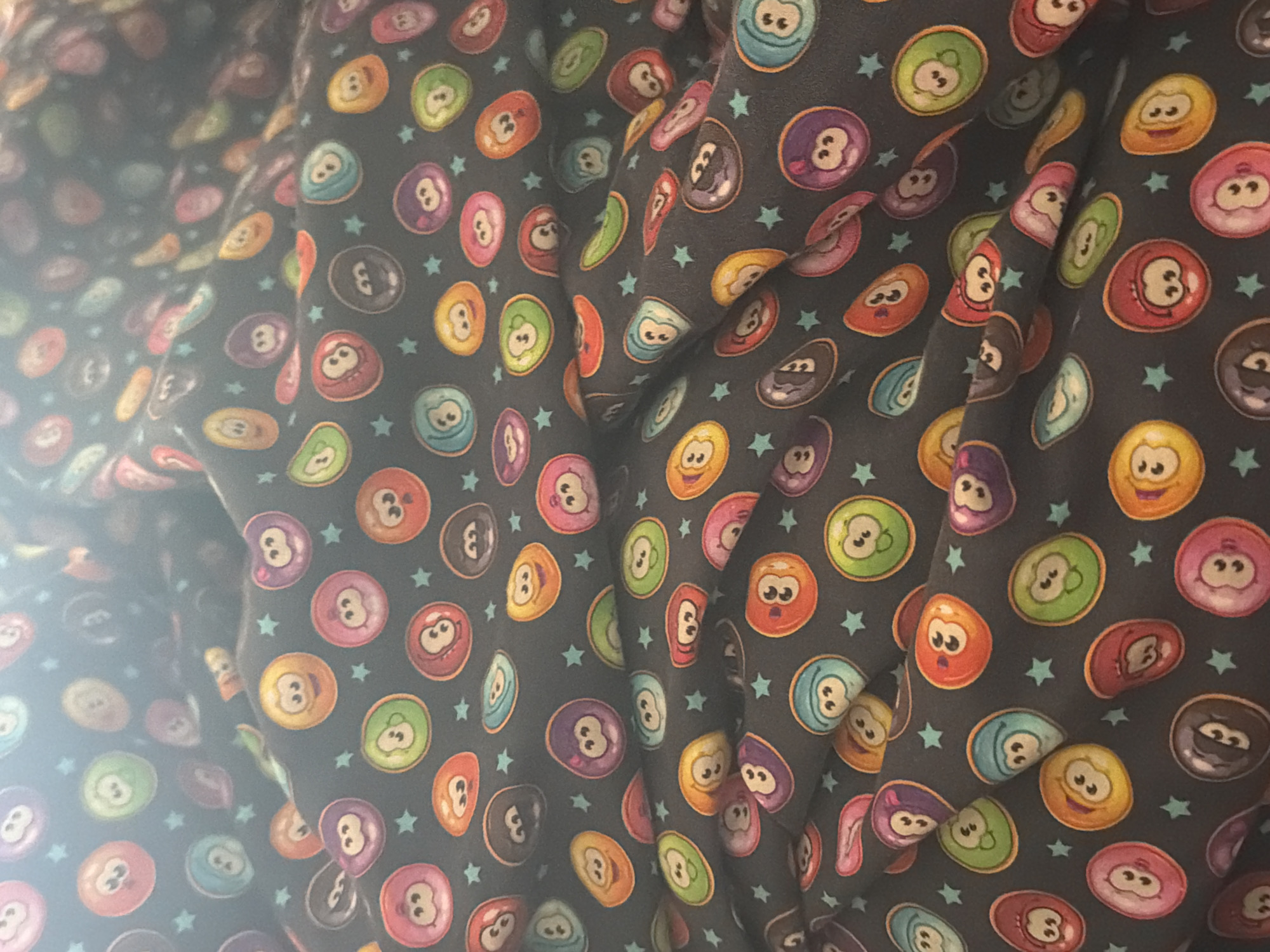 The image displays a close-up view of a dark-colored fabric featuring a vibrant, repeating pattern. The pattern consists of numerous irregularly shaped cartoon characters, resembling anthropomorphic billiard balls, set against a dark grey or black background. These characters are rendered in a variety of bright colors including red, orange, yellow, green, blue, purple, pink, and brown. Many of the characters have distinct facial expressions, ranging from happy and smiling to surprised, sleepy, or grumpy, and some also display the numbers '8' or '3' as part of their design. Small, light blue five-pointed stars are scattered across the fabric's background, interspersed among the colorful characters.
The fabric is visibly draped and folded, creating a textured surface with contours, shadows, and bright highlights. A strong light source from the left side of the frame illuminates the material, producing a prominent highlight and a slight lens flare effect, particularly on the left. The folds in the fabric partially obscure some of the pattern elements.
No specific activity or event is depicted; the image is a static portrayal of the patterned textile itself. There are no discernible location cues present within the image, nor any external text beyond the integrated numbers '3' and '8' on some characters. The material appears soft, suggesting it could be used for clothing, bedding, or other textile applications.