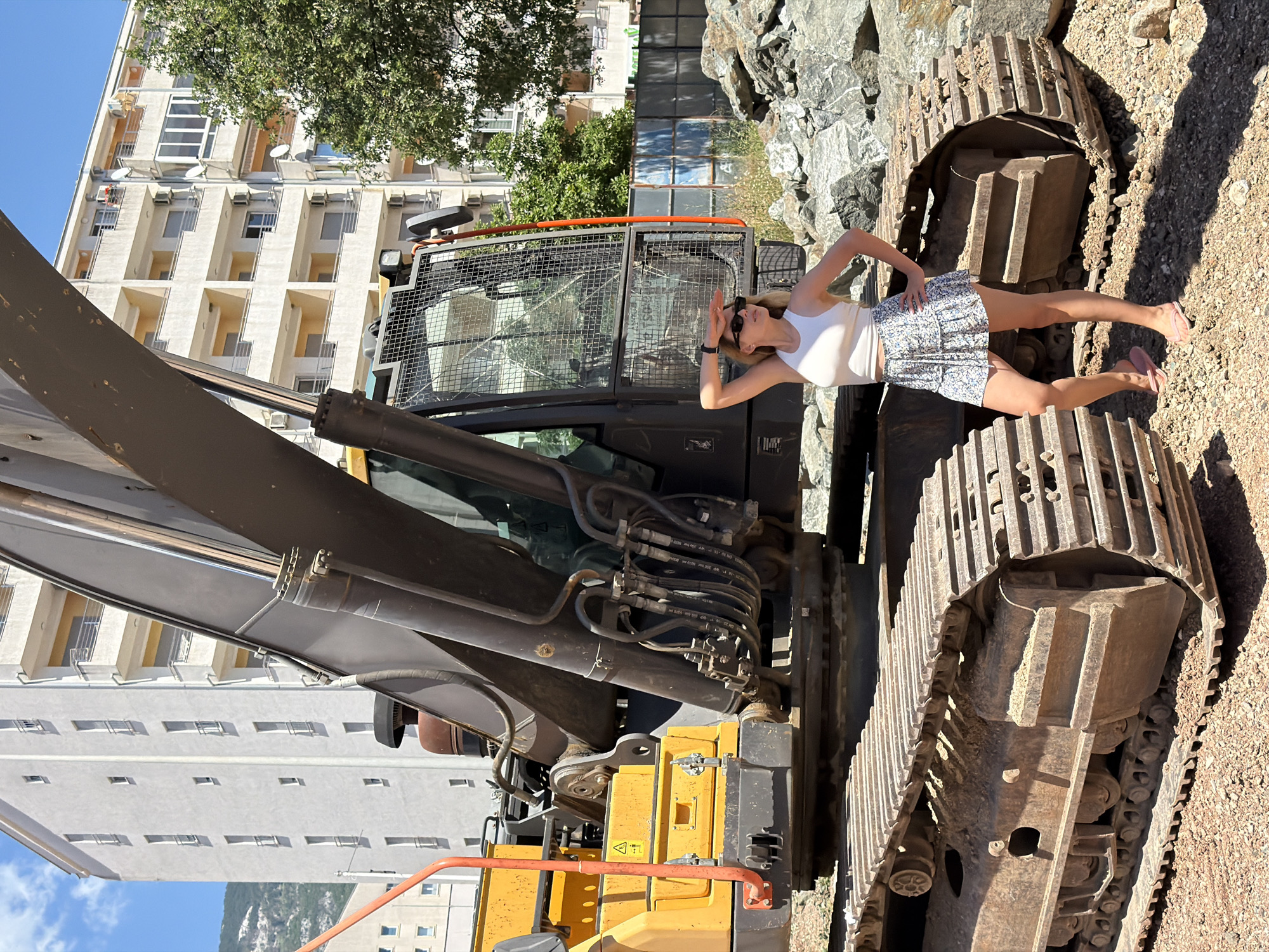 A young woman stands with her hand shielding her eyes from the sun, looking into the distance. She is wearing a white tank top and a floral-patterned skirt, along with pink flip-flops. The woman is positioned in front of a large yellow and black excavator, which is parked on a construction site with piles of rocks and dirt. In the background, there are several multi-story apartment buildings, suggesting an urban or suburban setting. The scene appears to be in the city of Budva, Montenegro, based on the architecture and the mountainous landscape visible in the far background. The sky is clear and blue.