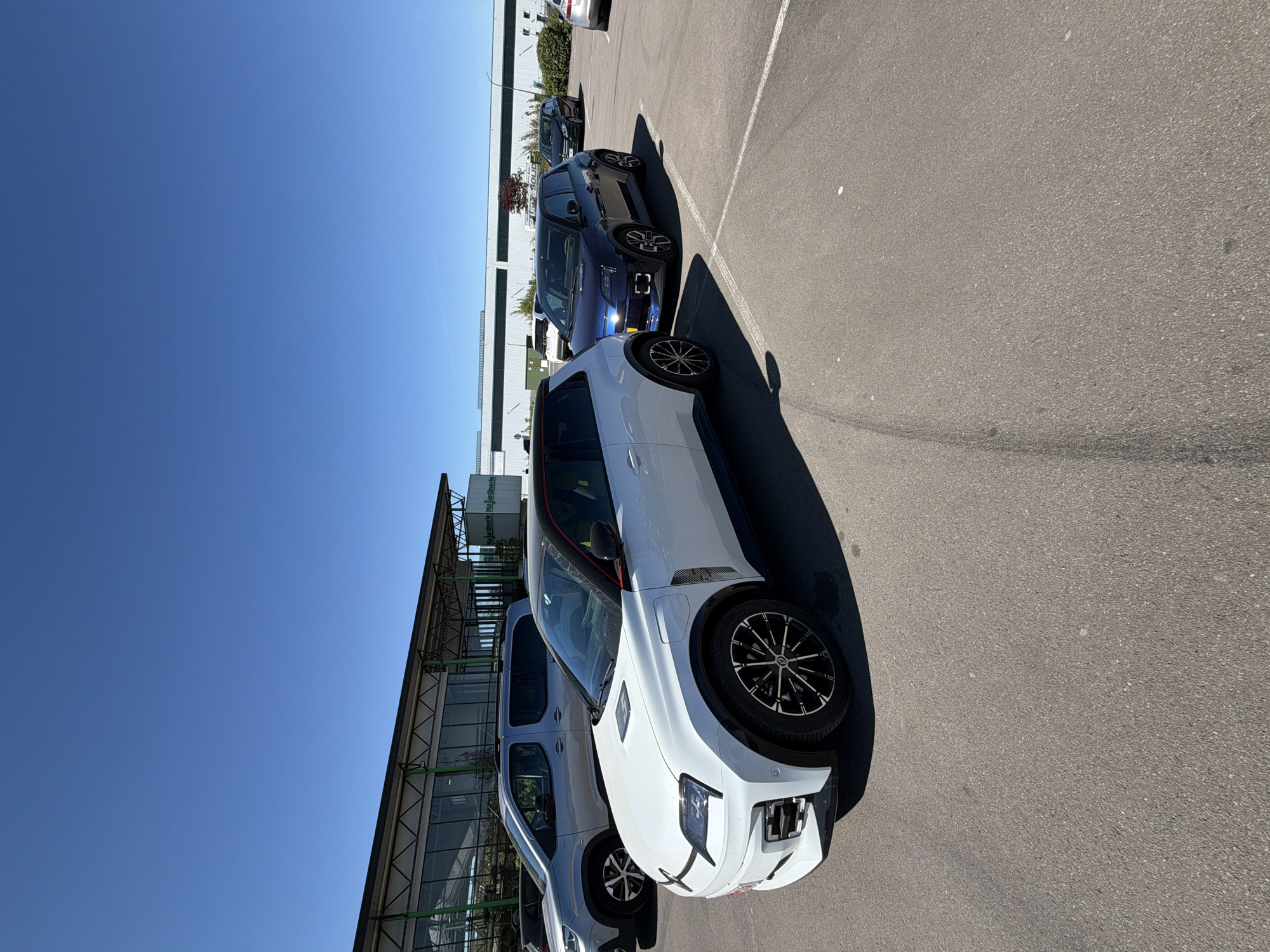 The image captures an outdoor parking lot under a bright, clear blue sky, likely in Aubange, Belgium. Several cars are parked on a gray asphalt surface marked with white lines.
In the foreground, a modern white SUV or crossover with black accents is prominently featured. It has distinctive black and silver multi-spoke alloy wheels, black wheel arch trim, and sharp, contemporary front lighting. A subtle red trim is visible along its roofline. Behind this vehicle, a dark blue car, possibly another SUV or a large hatchback, is parked, with its front lights illuminated. Further back, other cars are partially visible.
To the left of the parking area, a modern building with large glass panels and a visible metal framework extends across the scene. Some greenery, including bushes and small trees, lines the base of the building. The strong sunlight indicates it is daytime, likely midday or early afternoon, casting distinct shadows from the cars onto the ground. The sky is entirely cloudless.
On a white sign located on the building in the background, faint text that appears to read "jardinerie" is visible, suggesting the building may be a garden center or part of a commercial complex. No people are present, and the primary activity depicted is the parking of vehicles.