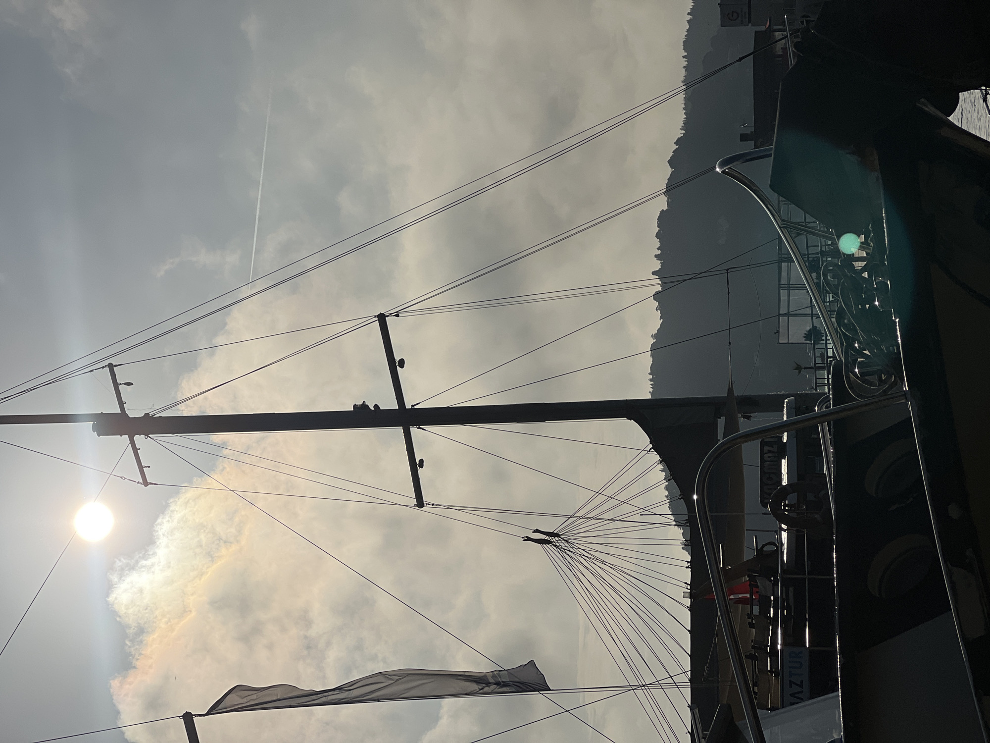 The image captures a silhouetted sailboat mast and rigging against a hazy sky with the sun partially obscured by clouds. The sun creates a bright halo effect. A flag is visible hanging from the left side of the mast. The foreground shows the upper deck of another boat with metallic railings and what appears to be a steering wheel. The name "MAZTUR" is visible on the side of this boat. In the background, a tree-covered hill or mountain range forms a silhouette against the sky, suggesting a coastal or waterfront location. The overall atmosphere is slightly overcast and diffused light. Given the context of Beşiktaş, Turkey, this scene likely depicts a harbor or marina with traditional Turkish Gulet-style boats, common in the region.
