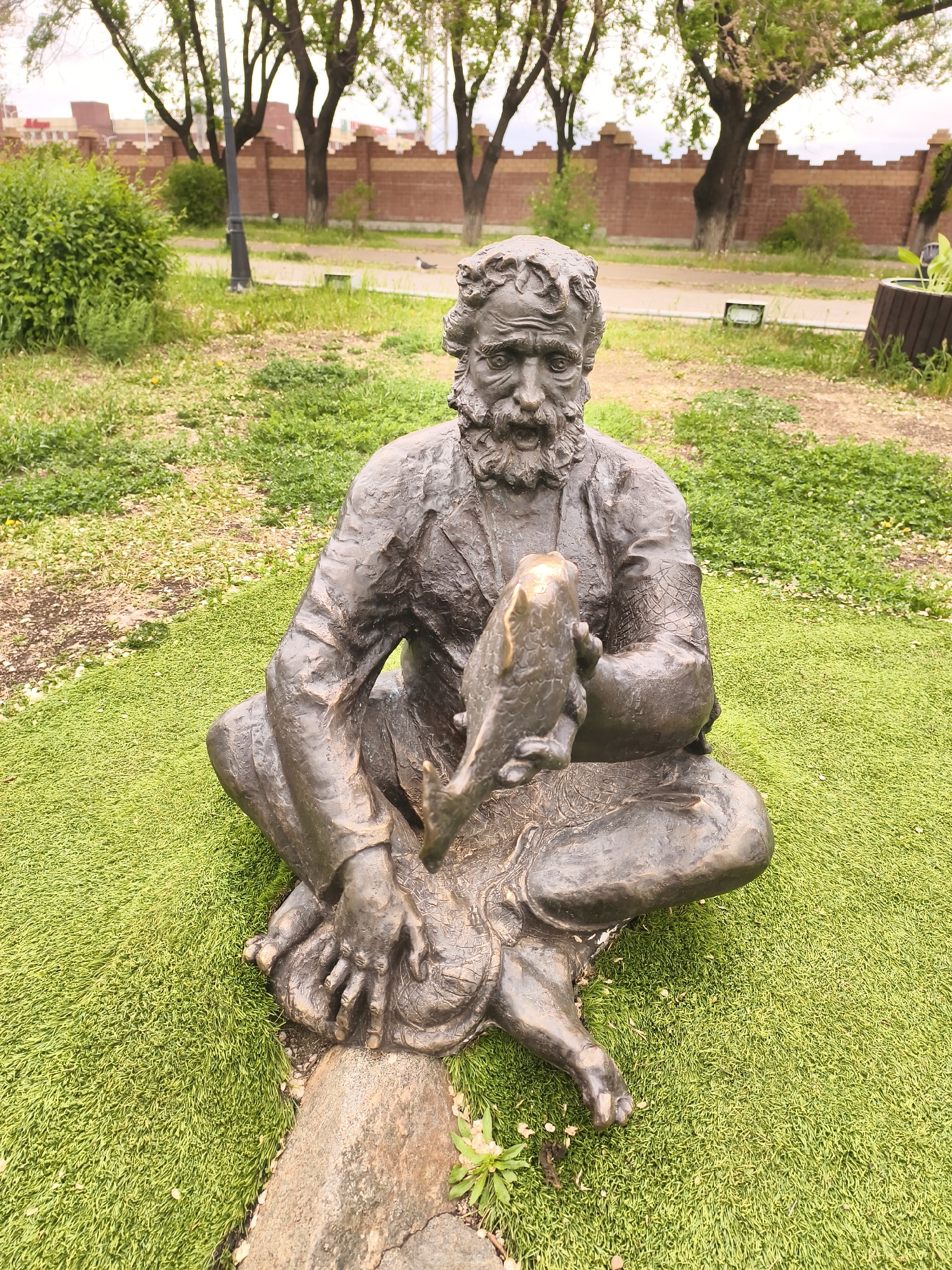 The image shows a statue of a man sitting cross-legged on a patch of green. The statue appears to be made of bronze. The man has a beard and is holding a fish in his hands. In the background, there are trees, a brick wall, and a pathway. The statue is located in the city of Blagoveshchensk, Russia.