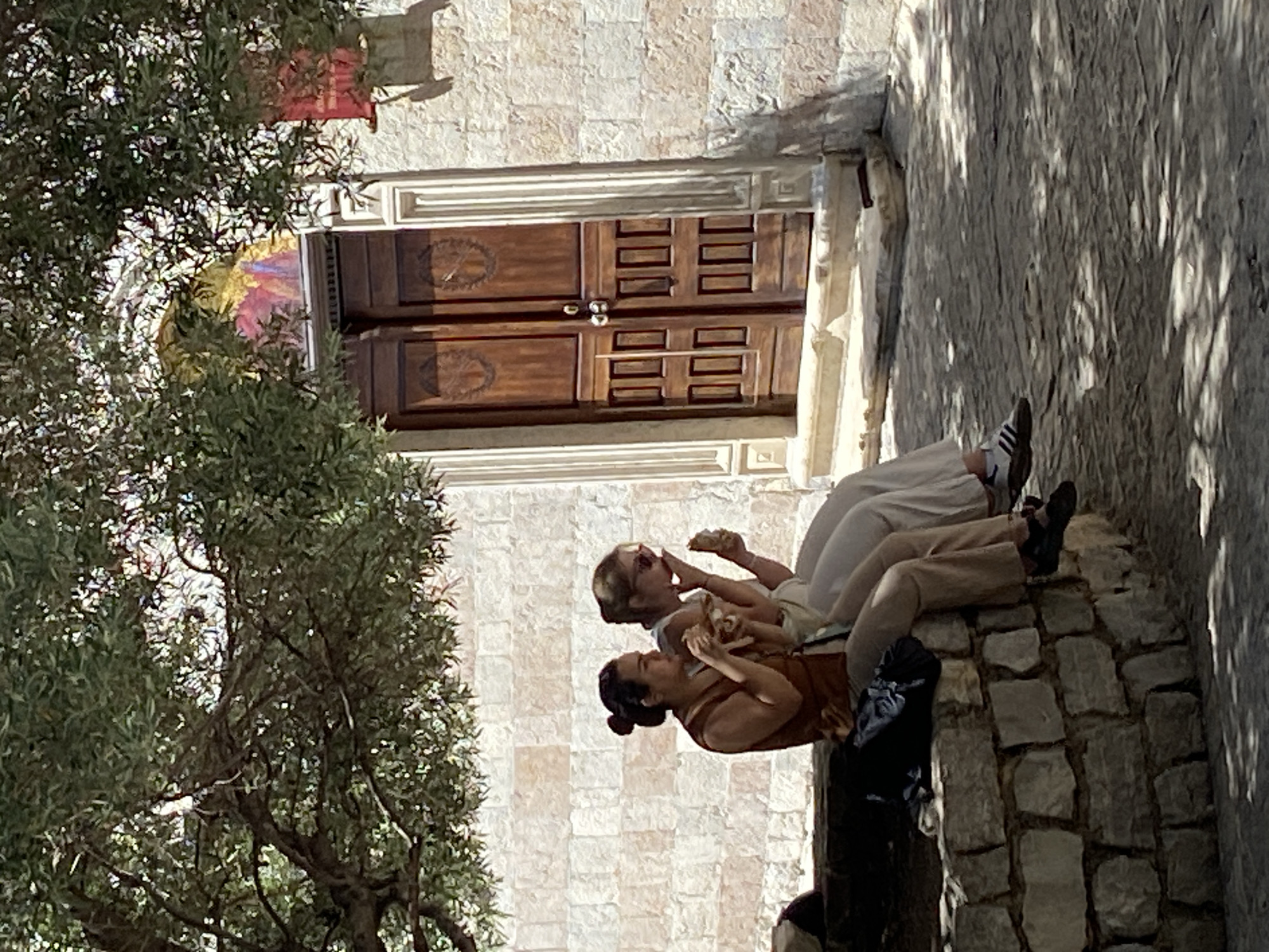 Two adult females are positioned in profile, seated on a low, curved stone wall, consuming food items, within the city of Budva, Montenegro. The subject on the left, with dark hair in a bun, wears a brown sleeveless top and light-colored trousers, holding a light-colored bread-based item. A dark bag rests on the wall next to her. The subject on the right wears sunglasses, a light-colored top, light-colored trousers, and white Adidas sneakers with black stripes; she is also eating a light-colored food item. They are situated beneath the dappled shade of a large, leafy tree whose branches extend into the upper frame. In the background, a building with light-toned, rectangular stone block masonry features a prominent double wooden door. This door is deeply paneled with carved circular motifs at the top and rectangular panels below, framed by decorative fluted pilasters. Above the door, a partially obscured, multi-colored fresco depicting figures is visible. A red rectangular sign is mounted on the stone wall to the right of the door. The ground surface is paved, exhibiting sharp contrasts of sunlight and shadow cast by the overhead foliage.