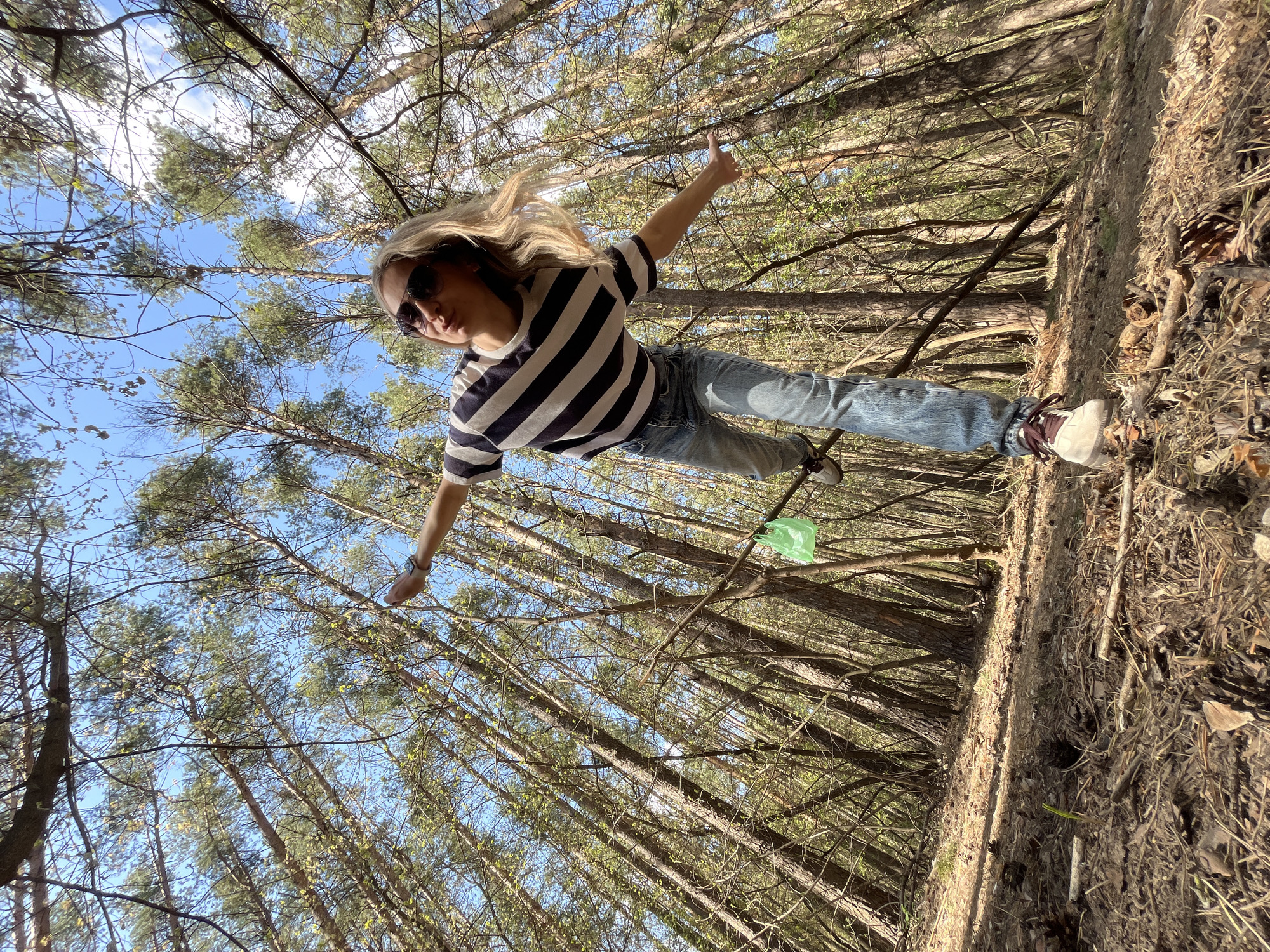 A woman stands on a fallen tree in a forest, near Chaikovsky, Russia. The photo has a low-angle perspective, looking up at the woman from the ground. She wears sunglasses, a striped short-sleeved shirt, jeans, and sneakers. Her arms are outstretched. The background consists of tall trees and a blue sky visible through the branches. A green plastic bag is caught on a tree branch. The ground is covered with dirt and fallen leaves.