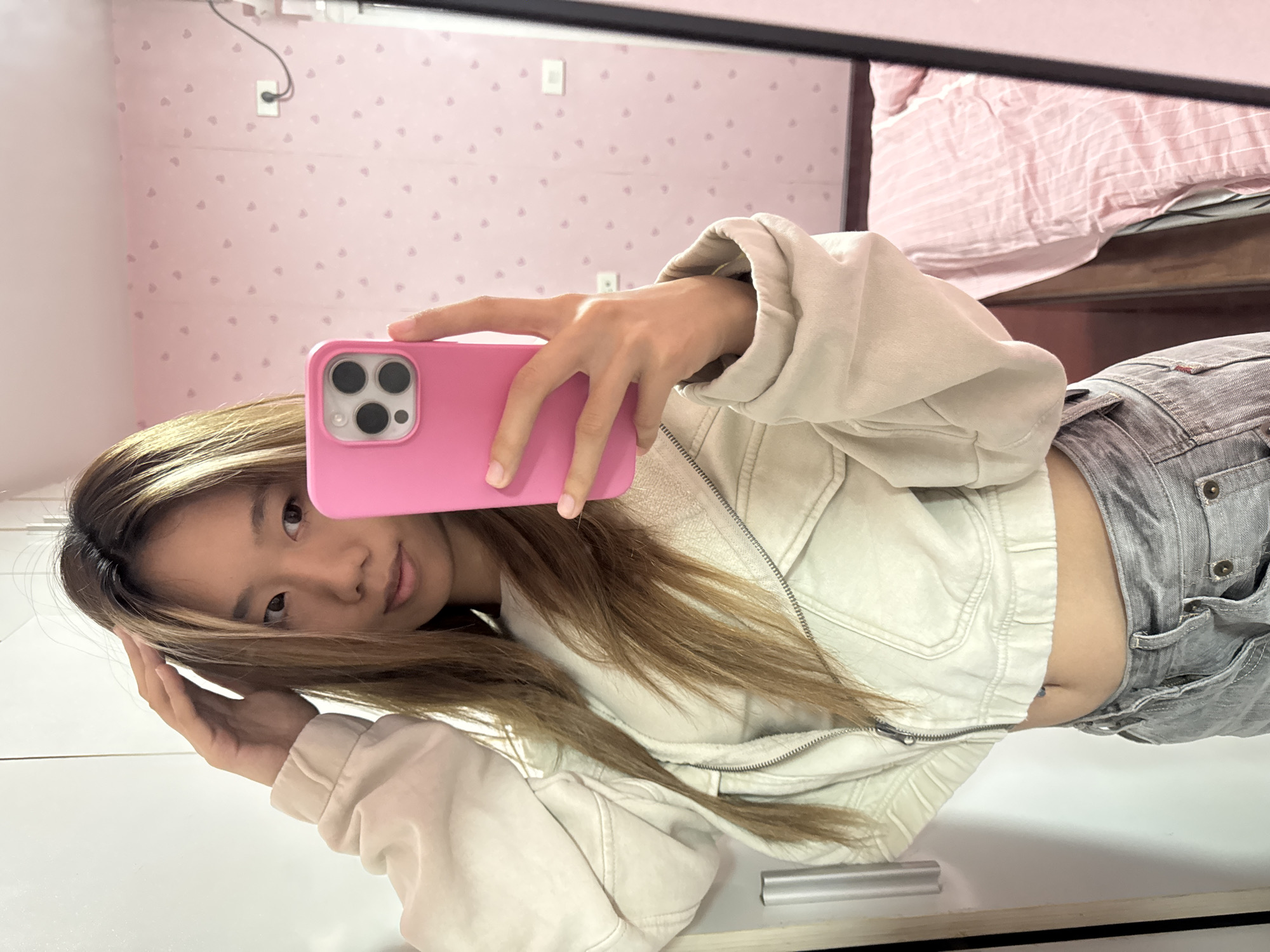 A young woman is taking a selfie in a mirror. She is holding a pink iPhone in her right hand. She has long, light brown hair and is wearing a white jacket with a zipper and gray pants. A glimpse of her midriff is visible. The background shows a pink wall with a small heart pattern and a bed with pink and white striped linens. The photo was probably taken indoors in a residential setting. It is not possible to confirm its location as Shapingba District, China without additional information.