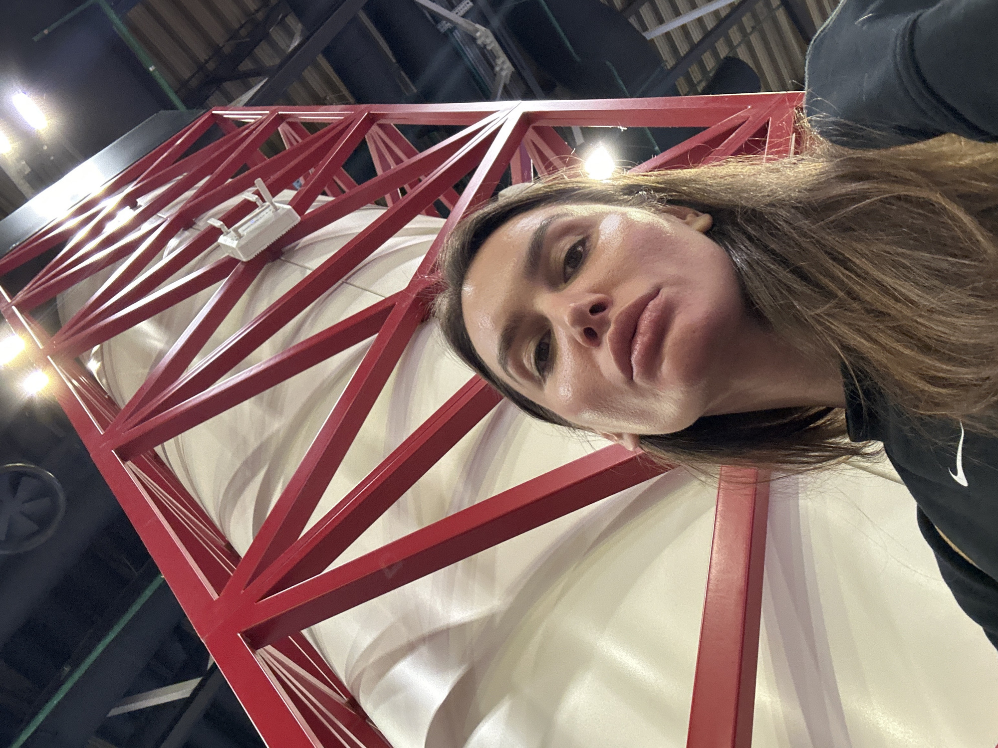A low-angle shot shows a woman in the foreground, with an industrial structure behind her. The structure is a red metal framework surrounding a large, white cylindrical object. The structure appears to be indoors, with ceiling fixtures visible in the background. A vent is visible on the left side of the frame. The woman is wearing a dark-colored garment with a white Nike logo. No specific details confirm the location as Лунёво, Russia.