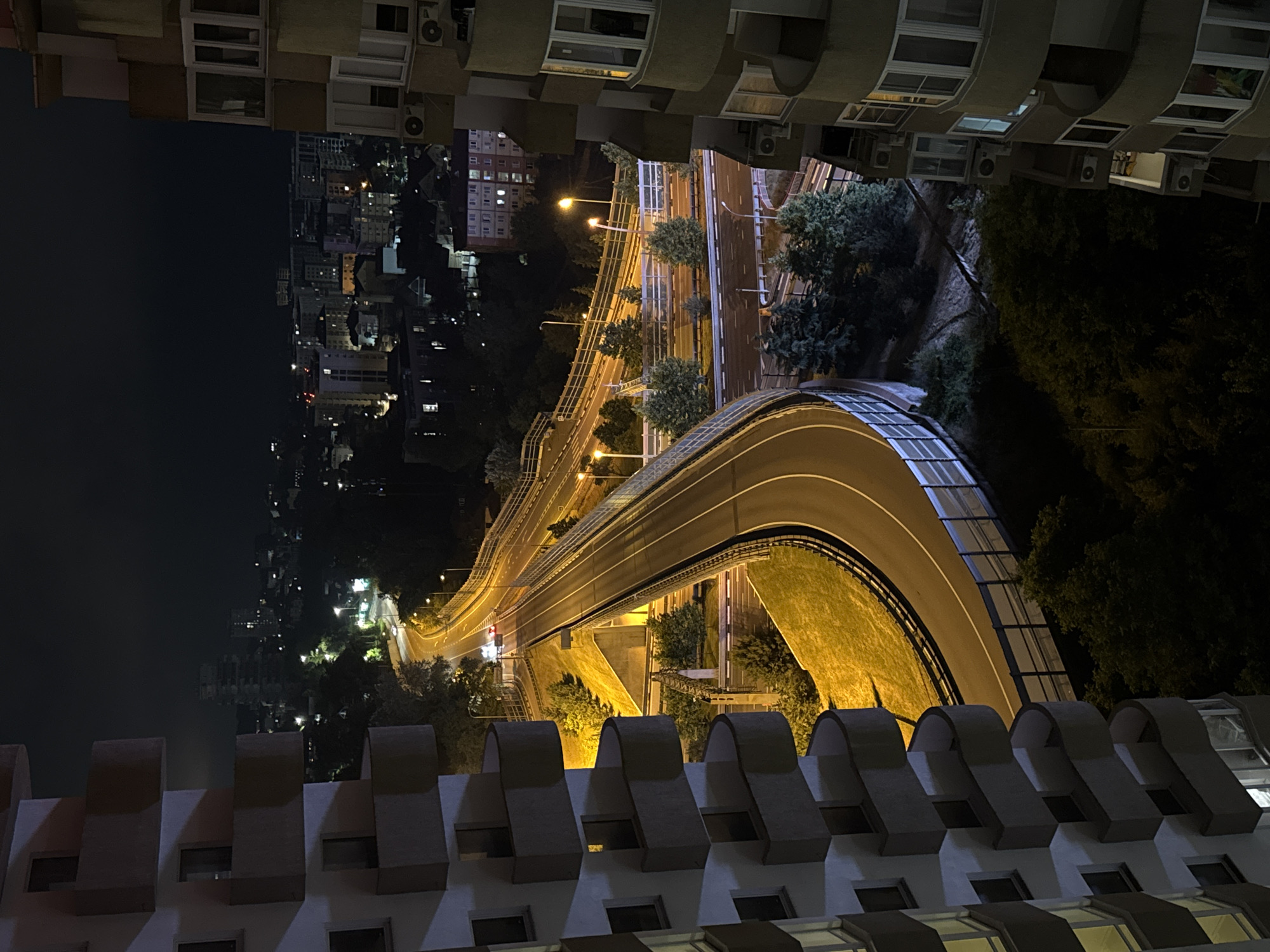 This is a nighttime, high-angle, long shot of a city in Sochi, Russia, featuring a highway overpass. On the left, a building with stacked, curved balconies and many windows is partially visible. On the right, another building with balconies and windows, along with air conditioning units, is also partially visible. The highway overpass, illuminated by streetlights, curves through the scene, with cars visible on it. Trees and other buildings are scattered throughout the background, with the city lights creating a glow against the dark sky.