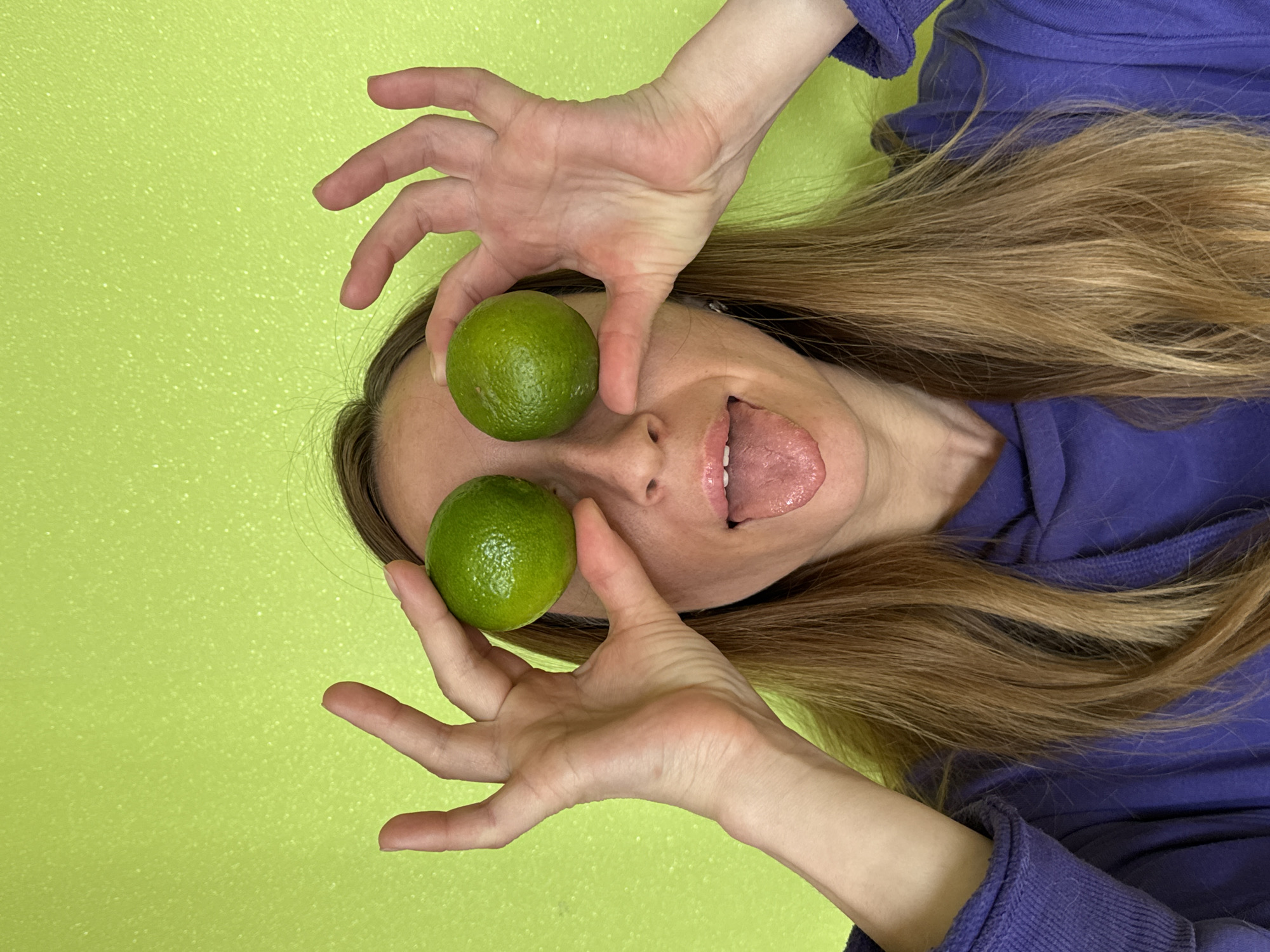 A person with long brown hair is positioned against a lime green background and a part of a blue hooded garment. The person's face is visible, with their tongue extended out. They are holding two green limes in front of their eyes as if they are replacing their eyes. The picture's orientation is rotated. The location in the city of Прикамский, Russia, is not evident.