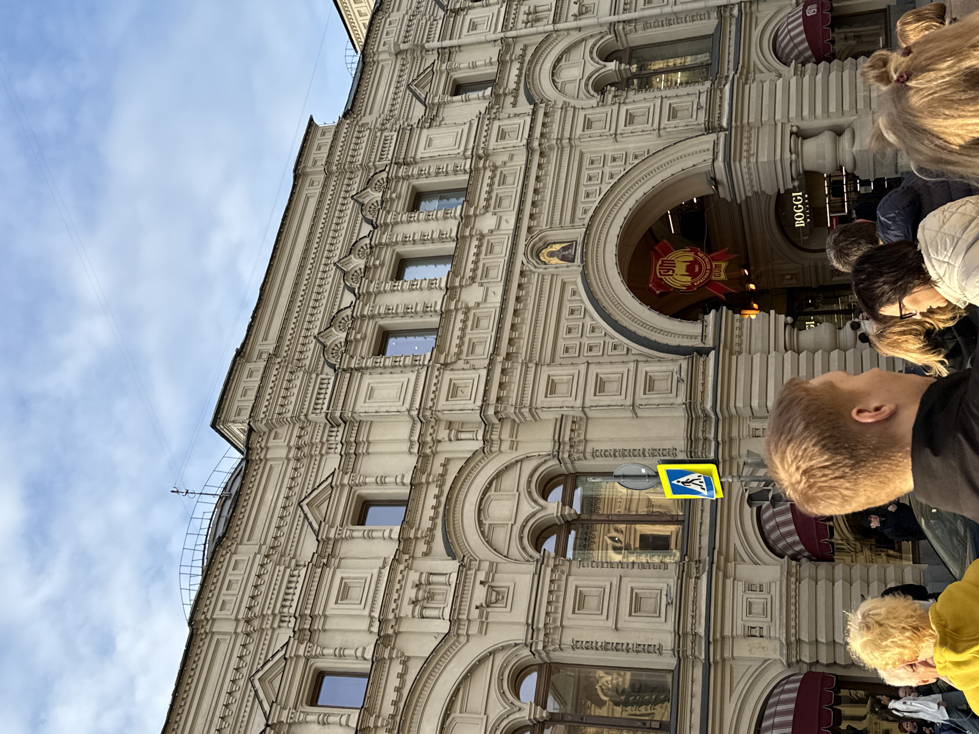 The scene is located in the city of Lyubertsy, Russia.
The image depicts a grand, multi-story building with ornate, light-colored architecture, likely stone or stucco, under a partly cloudy sky. The building features numerous decorative elements, including pilasters, cornices, and recessed rectangular and arched panels. On the lower levels, several large arched windows with brown frames are visible, some reflecting the surrounding environment. Above these, multiple rectangular windows are set into the facade. A prominent arched entrance is central, beneath which a large red banner with yellow text displays "ГУМ," "КРАСНАЯ ПЛОЩАДЬ" (Red Square), and "GUM." Below this banner, a sign for "BOGGI MILANO" is visible within the archway. Directly above the main arch, a small, golden-hued icon of a religious figure is embedded in the facade. On the roofline, a metal railing partially encircles a curved structure, possibly a skylight or dome, with utility lines stretching across the sky.
In the foreground, a group of people is present, viewed mostly from behind or the side. A person with short, reddish-blonde hair in a dark jacket occupies the immediate right foreground. To their left, a person with bright blonde, curly hair wears a yellow hooded garment. Other individuals are partially visible, including a woman with dark hair and glasses in a light quilted jacket. They appear to be pedestrians on a street or sidewalk, oriented towards the building. A blue square sign with a white pedestrian symbol, indicating a crossing, is mounted on a dark pole in the mid-ground. Red and white striped awnings are extended over some ground-level windows or entrances to the left of the main archway.