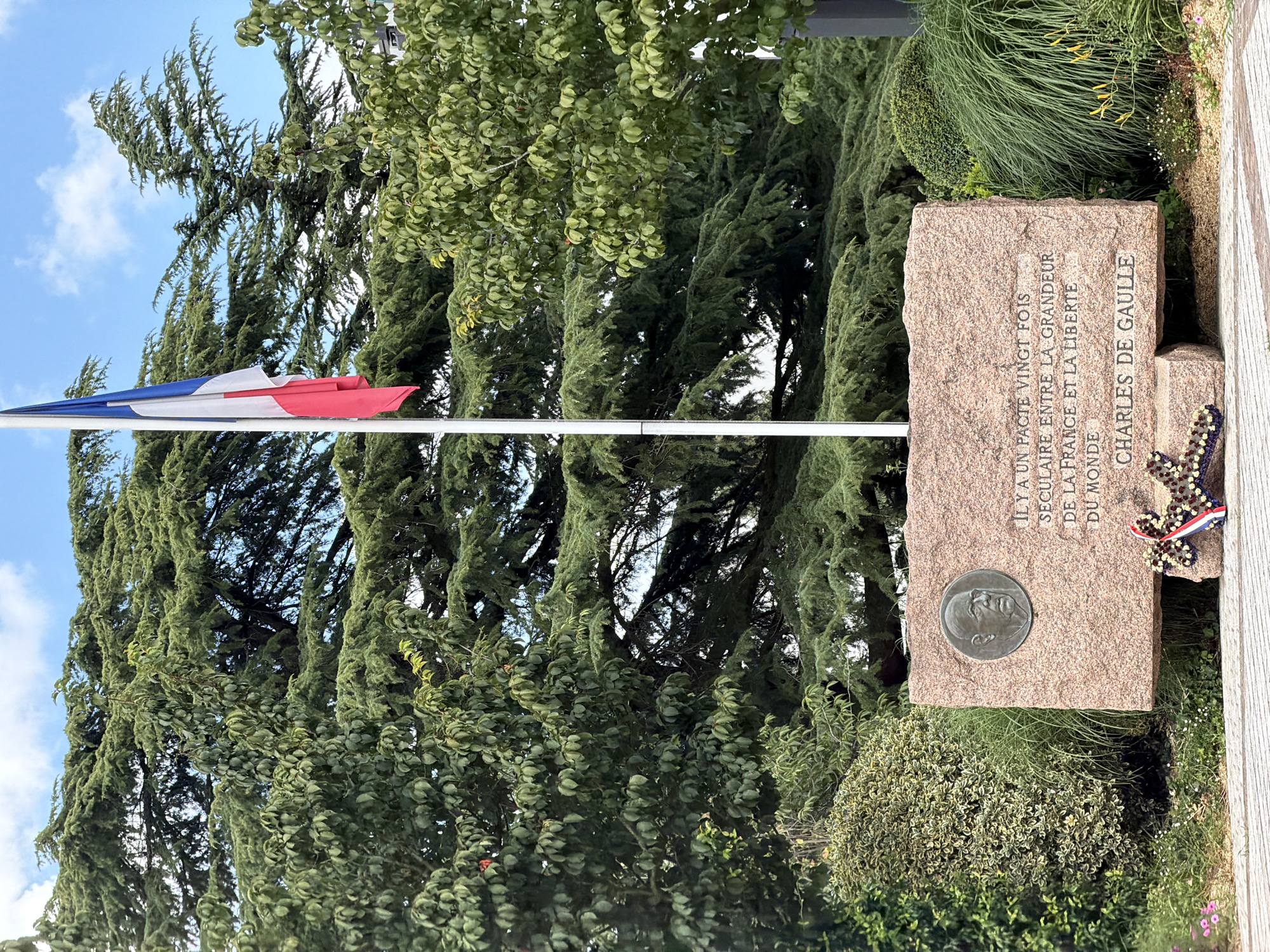 This media file shows a monument dedicated to Charles de Gaulle, situated outdoors amidst lush greenery. The monument itself is a large, rough-hewn stone, likely granite, with an engraved plaque and a circular bronze medallion featuring a portrait of de Gaulle. The engraved text reads: "ILY A UN PACTE VINGT FOIS SECULAIRE ENTRE LA GRANDEUR DE LA FRANCE ET LA LIBERTE DU MONDE CHARLES DE GAULLE." A wreath adorned with the French tricolor flag and a pattern of dark beads is affixed to the side of the monument.
To the left of the monument, a flagpole flies the French flag, which appears to be slightly furled. The background is dominated by dense foliage from trees, with some sunlight filtering through, suggesting it is daytime. The sky is visible in the upper left quadrant, with some clouds present. The overall scene evokes a sense of solemnity and remembrance, likely a memorial site or a public square commemorating a significant historical figure. The presence of the French flag and the text on the monument clearly indicate a French context, and the location is identified as Vélizy-Villacoublay, France.