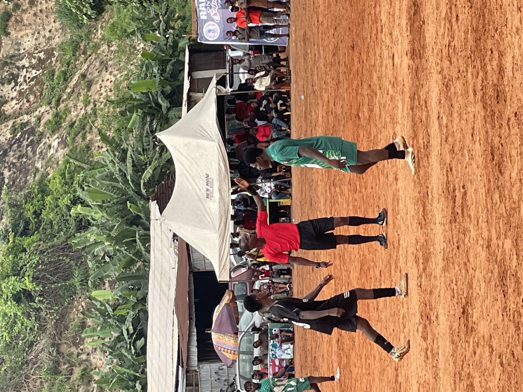 In a low-angle, full shot, a soccer match is underway on a dirt field under an overcast sky. A referee in a red shirt and black shorts stands in the center of the frame, facing right with his left arm raised. Two young male soccer players, one in a black jersey with white stripes and black shorts, and the other in a green jersey and shorts, are positioned on the field. In the background, spectators are gathered behind a barrier under tents and a canopy. To the left, an umbrella shades a group of people and vehicles. Lush green vegetation covers a hillside in the upper background, with some signs and banners visible. The ground is made of red dirt, indicative of a typical sports field in many regions. The overall scene depicts a casual, local sporting event.