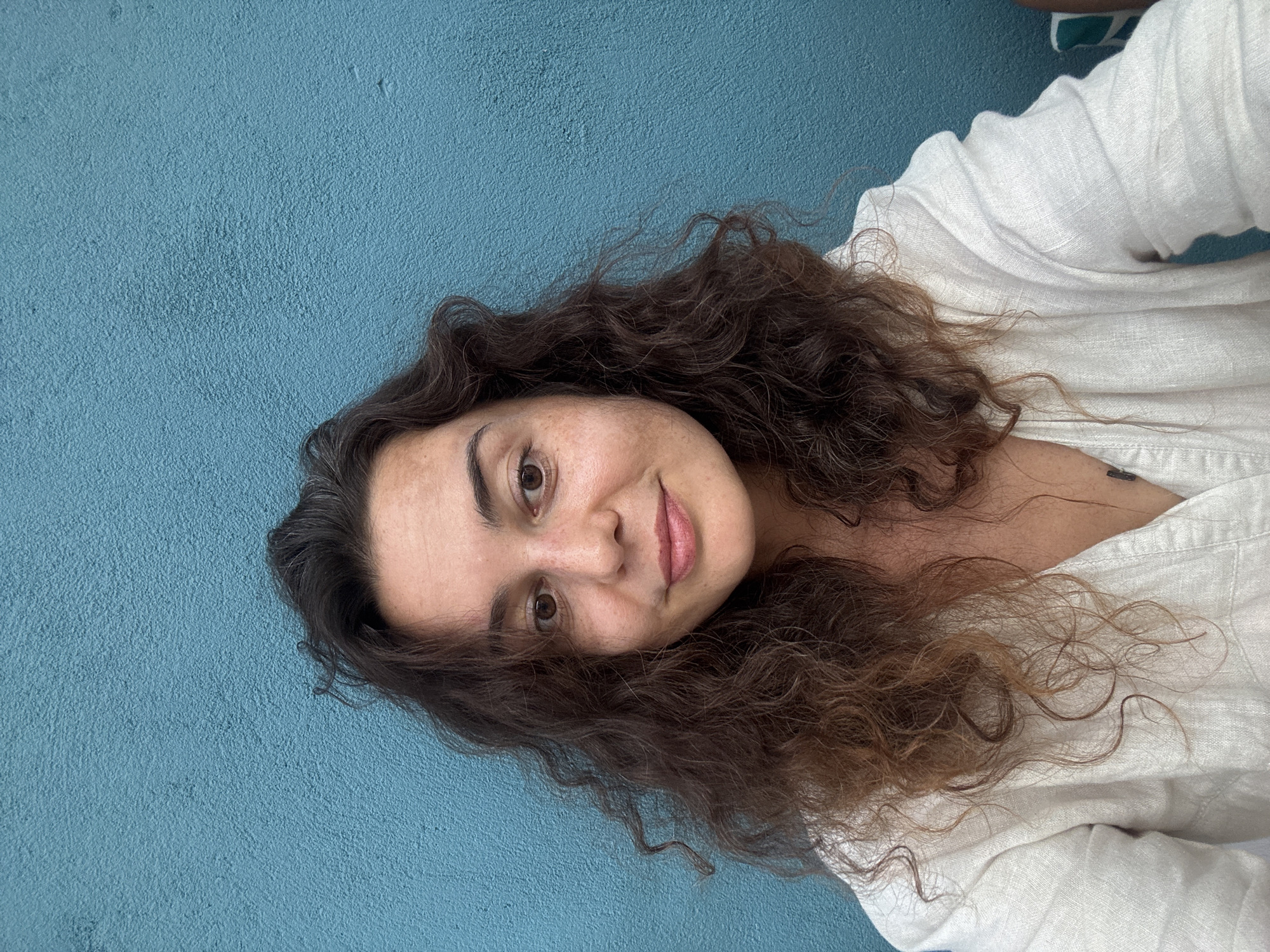 A woman with curly brown hair is pictured from the chest up, looking at the camera. She is wearing a light-colored, collared shirt with a V-neck. The background is a textured, bright blue wall. The image appears to be taken outdoors, possibly in the city of Hoi An, Vietnam, given the characteristic textures and colors often found in that region. There are no other people or significant objects in the frame.