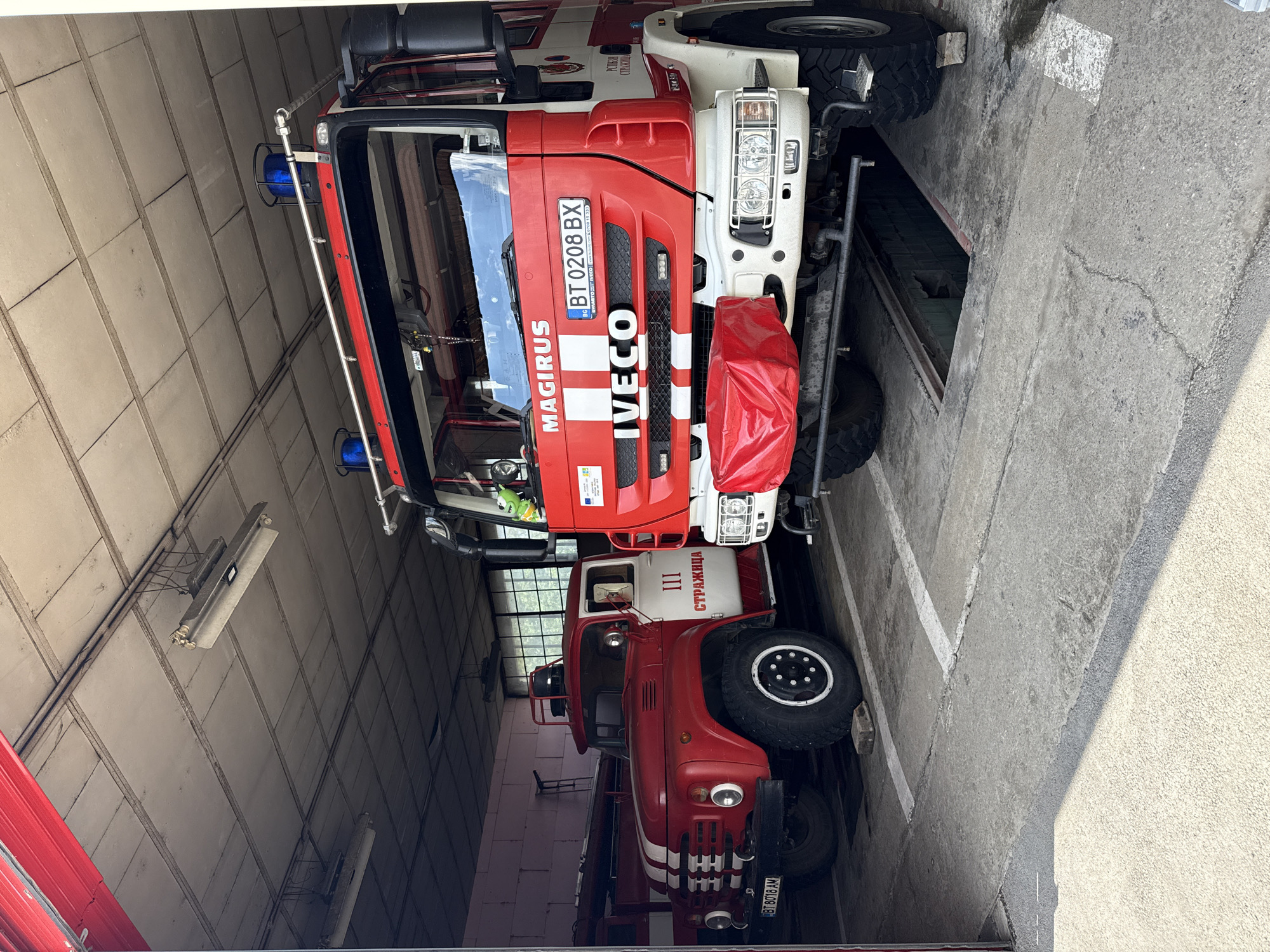The image shows two fire trucks parked in a garage in Strazhitsa, Bulgaria. The first fire truck is a newer model, red and white, with the word "MAGIRUS" and "IVECO" visible. It has a license plate that reads "BT 0208 BX". The second fire truck is an older model, primarily red, with "III СТРАЖИЦА" written on its white panel. Its license plate reads "BT 8018 AM". The garage has a concrete floor and ceiling. The image is taken from the entrance of the garage, looking inside. Fluorescent lights are visible on the ceiling.