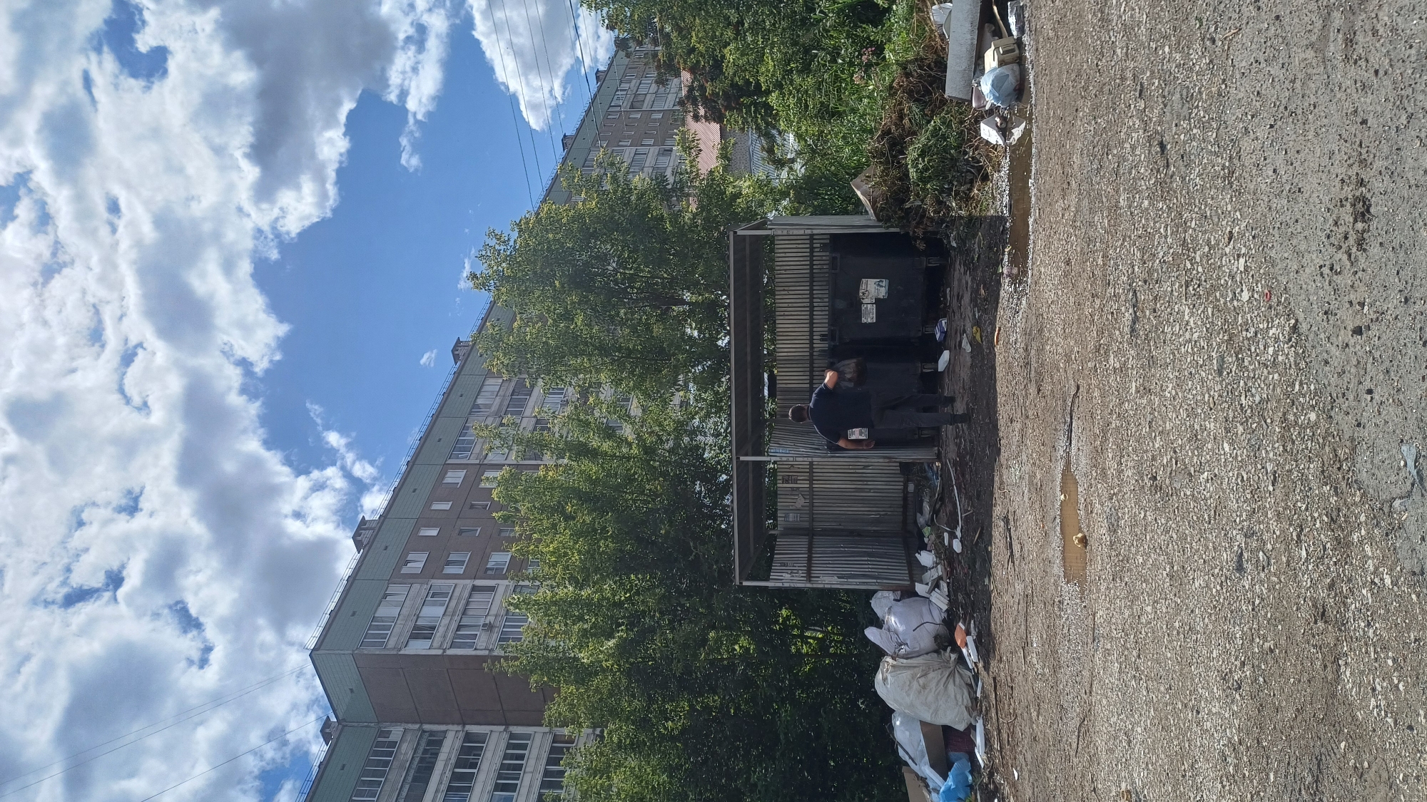 The image depicts an outdoor scene at a garbage collection point in Izhevsk, Russia. In the background, a multi-story apartment building, typical of Soviet-era architecture, dominates the skyline. The building is light brown with green trim and features numerous windows. In front of the building, a row of mature trees forms a dense green barrier.
The foreground is a cluttered area around a metal shelter housing large black garbage bins. One man, dressed in a dark shirt and trousers, is seen from behind, interacting with one of the garbage bins. He appears to be either disposing of trash or retrieving something. Piles of garbage bags and scattered debris surround the shelter and the bins, indicating a significant amount of waste accumulation. The ground is uneven and appears to be a mixture of dirt and gravel, with a noticeable puddle of water near the center of the frame. The overall impression is one of disarray and neglect at a common waste disposal site. The sky above is blue with scattered white clouds.