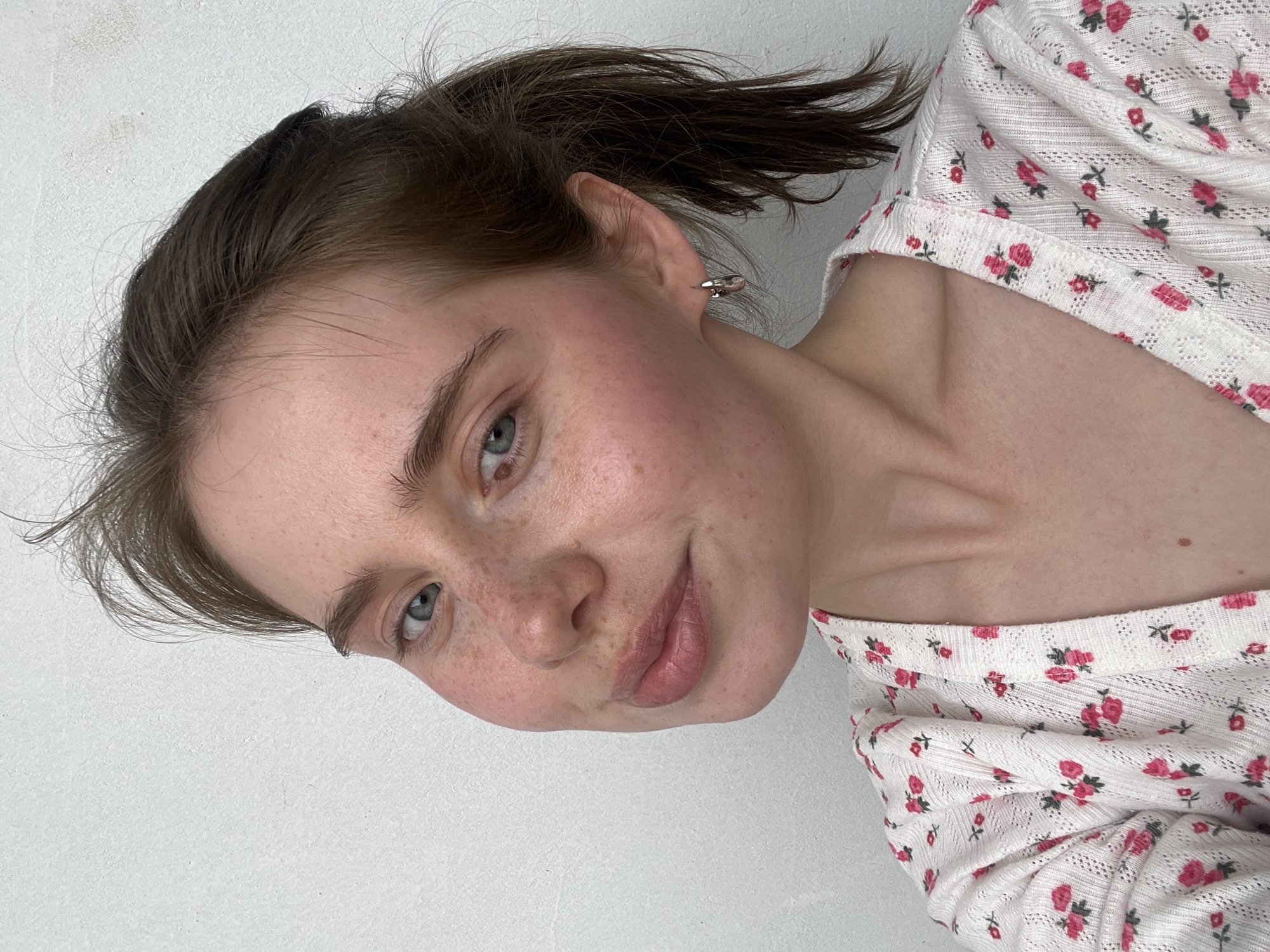 A close-up, slightly rotated portrait shows a young woman with fair skin, freckles, and light brown hair pulled back in a ponytail. She has light blue eyes, slightly rosy cheeks, and a faint smile. She wears a white, floral-patterned top with small pink roses. A silver earring is visible on her left ear. The background is a plain white wall. It is not possible to determine if the location is in Chaikovsky, Russia based solely on this image.
