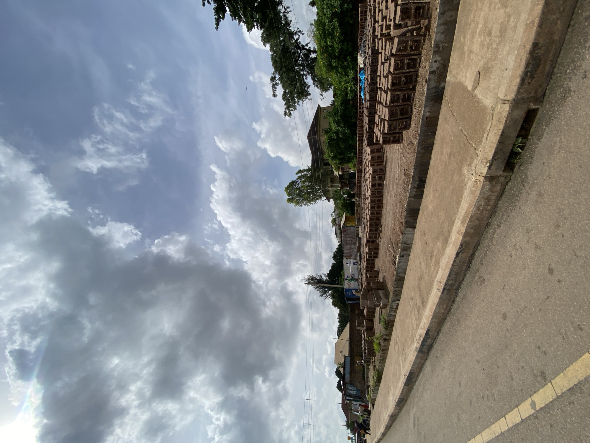 The image captures a scene in Jos, Nigeria, featuring a roadside area with a large quantity of bricks neatly stacked. The foreground shows a paved road with a yellow line marking. Adjacent to the road is a concrete walkway and a raised curb. The main subject of the image is the extensive array of bricks, organized in rows, suggesting a brick-making or storage facility. In the background, several buildings are visible, including what appears to be a residential house with a brown roof, and other smaller structures. Trees and lush green vegetation surround the buildings. The sky is filled with dramatic, textured clouds, indicating dynamic weather conditions. A few individuals can be seen in the distance, seemingly working or supervising the brick operation. The overall environment suggests an active, semi-industrial area within the city of Jos.