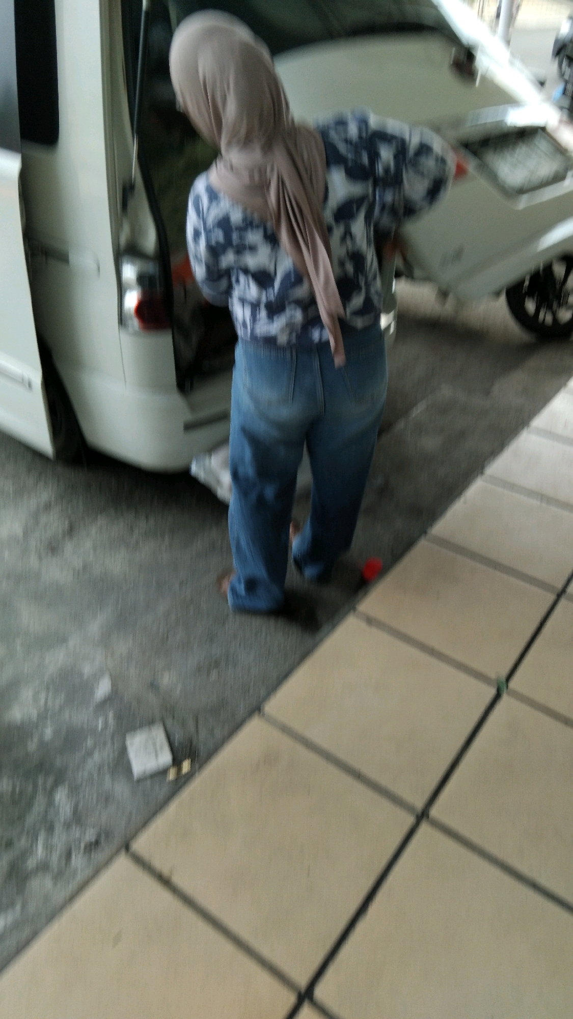 A person wearing a tan hijab and a patterned shirt with blue and white elements, along with blue jeans, stands with their back to the camera. They are positioned next to the open rear of a white van, with its doors ajar. The ground is a mix of rough concrete and tiled pavement. In the background, another white vehicle, possibly a different model of van or car, is partially visible with its wheel turned. There is a small red object on the tiled ground to the right of the person. The scene appears to be an outdoor area, possibly a parking lot or a service area, within the city of Pekalongan, Indonesia, given the context provided. The image is slightly blurred, suggesting motion or a low-quality capture.