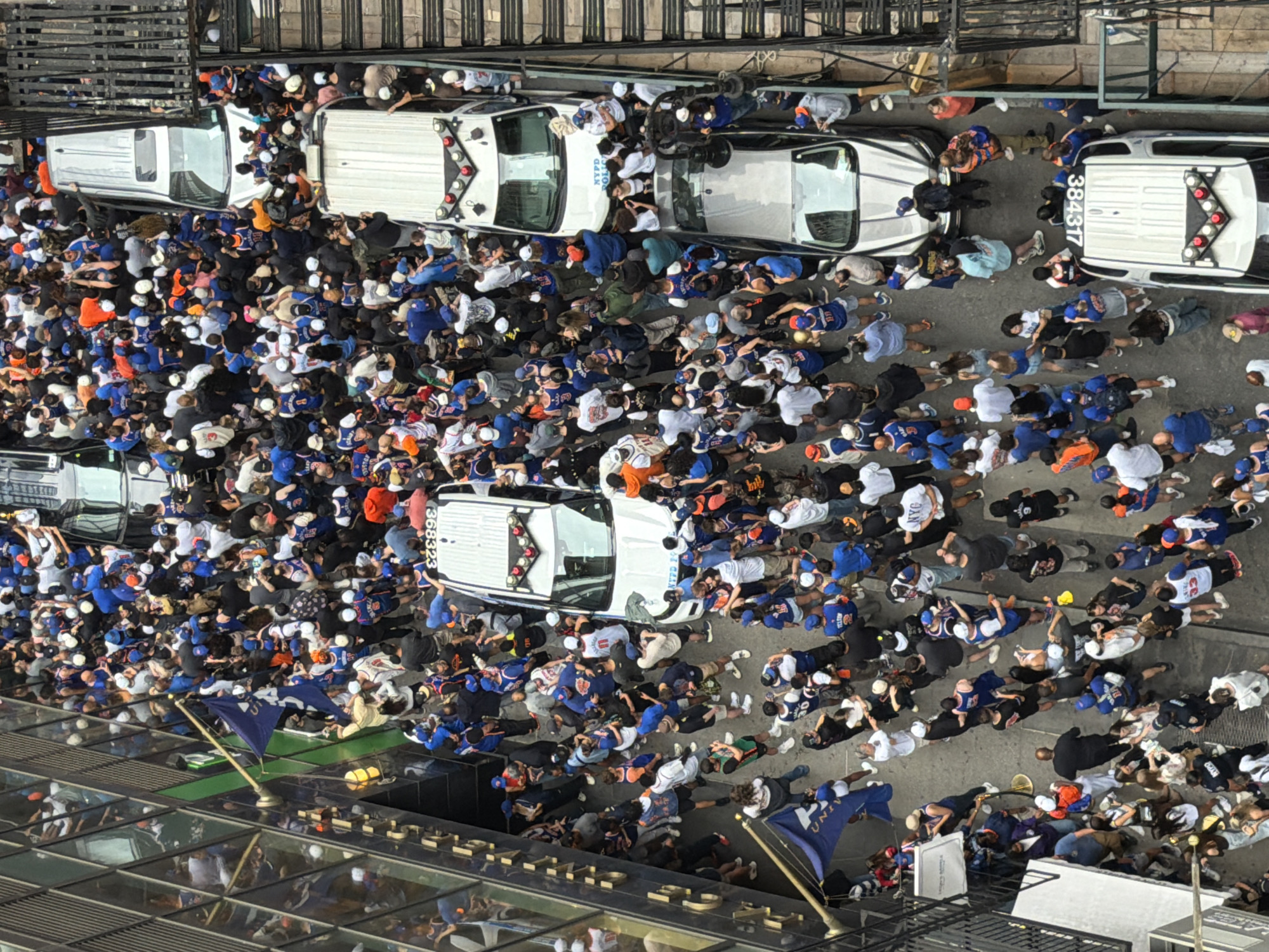 This aerial photograph captures a large crowd of people gathered on a street in New York City. The scene is dominated by a dense throng of individuals, many wearing blue and orange, suggesting they might be fans of a local sports team. They are clustered around several white vehicles, including what appear to be NYPD vehicles. The people are standing, some interacting with each other, while others are looking towards the vehicles or away from the camera.
The event taking place seems to be a public gathering or potentially a situation where the police are present due to the crowd. The density of the people and their attire suggests a lively event, possibly a celebration or a demonstration. The overhead perspective indicates the photo was taken from a significant height, perhaps from a building or a drone.
Notable details include the presence of what seems to be an NYPD vehicle, evidenced by "NYPD" visible on one of the vehicles. There is also text visible on the back of one of the white vehicles, reading "384317" and "368323". The lighting suggests it is daytime, possibly late afternoon or early evening. The weather appears to be clear, with no visible rain or snow. The overall atmosphere seems energetic due to the large number of people. In the foreground, a glass-covered walkway or building facade reflects the scene, adding another layer to the visual. A flag with blue fabric and a white emblem is visible, held by a pole.
