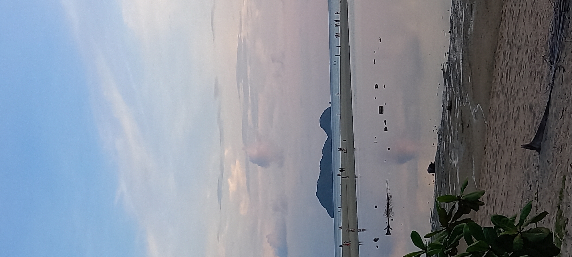 The image shows a coastal scene at Baan Sri Thanu, Thailand, featuring a long, narrow sandbar extending into the sea. The sky is partially cloudy. Several people are seen walking along the sandbar. Two islands are located on the sandbar that are close to one another. Reflections of these features are visible in the water. The right side of the image includes a sandy beach area with some vegetation in the foreground. Debris is also visible on the beach.