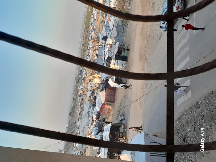 An elevated, downward-angled photograph, framed by dark, out-of-focus vertical bars, captures a sprawling displacement camp within Khan Yunis, Palestinian Territory, under a clear, pale blue sky. A central, unpaved roadway, marked with a white pedestrian crosswalk, traverses the mid-ground. On this road, a figure in dark clothing walks away, carrying a white bundle and a dark item, stepping onto the crosswalk. To the right, a silver sedan is parked, with a person in a red shirt standing nearby, and another individual partially visible beside the car. On the left side of the road, two individuals walk. The camp comprises a dense array of makeshift shelters constructed from white and light-colored tarpaulins and fabrics, extending across the landscape towards a distant horizon featuring low hills and sparse vegetation. A tall water tower is visible on the far left horizon. The ground throughout the camp is primarily dirt and sand, with scattered debris. Several other indistinct figures are visible moving between the shelters in the background.