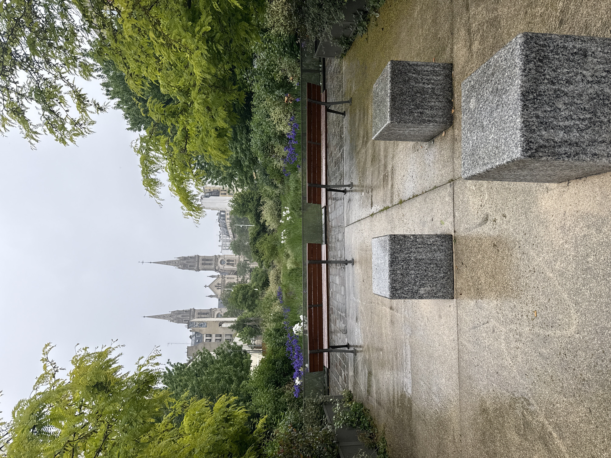 The image captures a serene, rainy day in Paris, France, presenting a cityscape viewed through a park setting. Two pointed towers of a Parisian church or cathedral rise above the greenery. In the foreground, a paved pathway leads the eye into the scene. Two wooden benches with metal frames are positioned along the pathway, accompanied by three gray, granite cube-shaped blocks. Lush green trees and various plants border the pathway. The overall atmosphere is quiet and damp, reflective of the rainy weather.