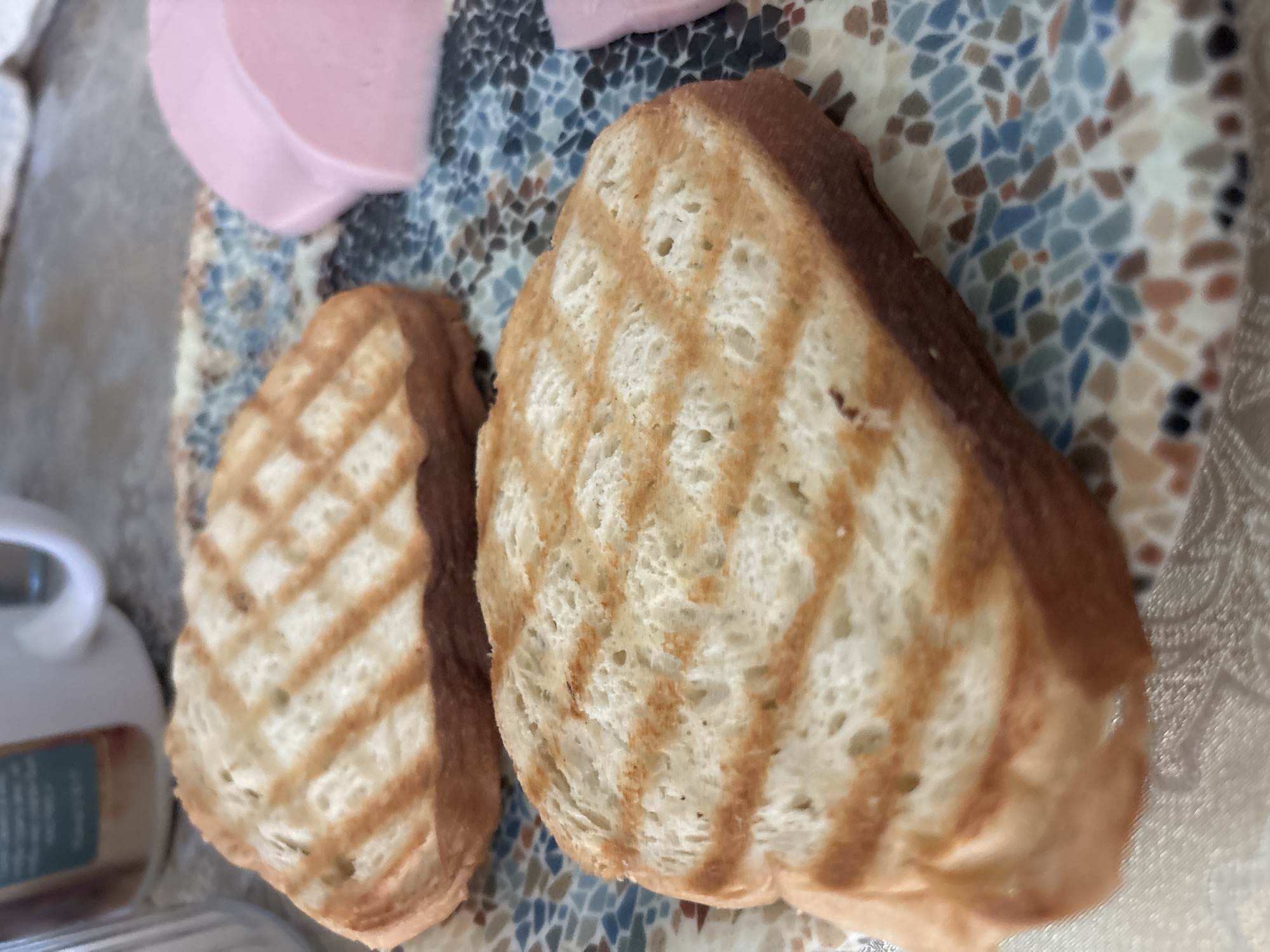 The image presents a close-up, high-angle view of two slices of grilled bread and several slices of deli meat, arranged on a decorative plate. The two main subjects are golden-brown bread slices, each displaying prominent, darker brown grill marks in a cross-hatch pattern, suggesting they were toasted on a griddle or in a panini press. The texture of the bread appears soft with visible air pockets.
In the upper left portion of the plate, several thin, round slices of light pink deli meat, likely ham or sausage, are partially visible. The plate itself features a distinctive mosaic or terrazzo-like pattern, composed of irregularly shaped pieces in various shades of blue, brown, beige, and off-white.
In the blurred background to the left, a white ceramic mug with a handle is partially visible, alongside a container with an indistinct label. The scene suggests a simple meal or snack, possibly breakfast, set indoors on what appears to be a table with a light-colored surface or tablecloth. The bright, natural lighting indicates it is daytime. No people are visible, and no discernible text provides specific information about the contents of the container. The overall impression is one of a straightforward, homely food presentation.