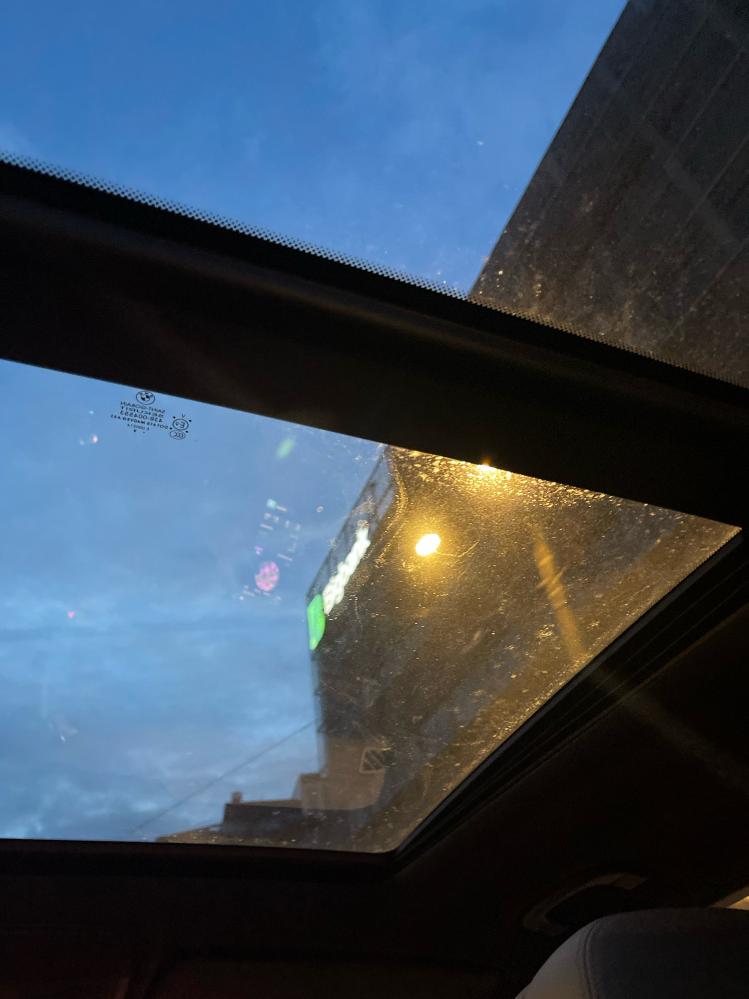 The image appears to be taken from inside a vehicle, looking up through the sunroof at a twilight sky. A building with a prominent green illuminated sign is visible in the distance, reflecting light onto the wet sunroof. The sign's text is unreadable due to the distance and reflections. The sky is a deep blue, suggesting either early morning or late evening. There are some smudges and water droplets on the sunroof, further obscuring the view. The interior of the car, including the headliner and a seat, is visible in the foreground. There is no indication of the location being Tallinn, Estonia.