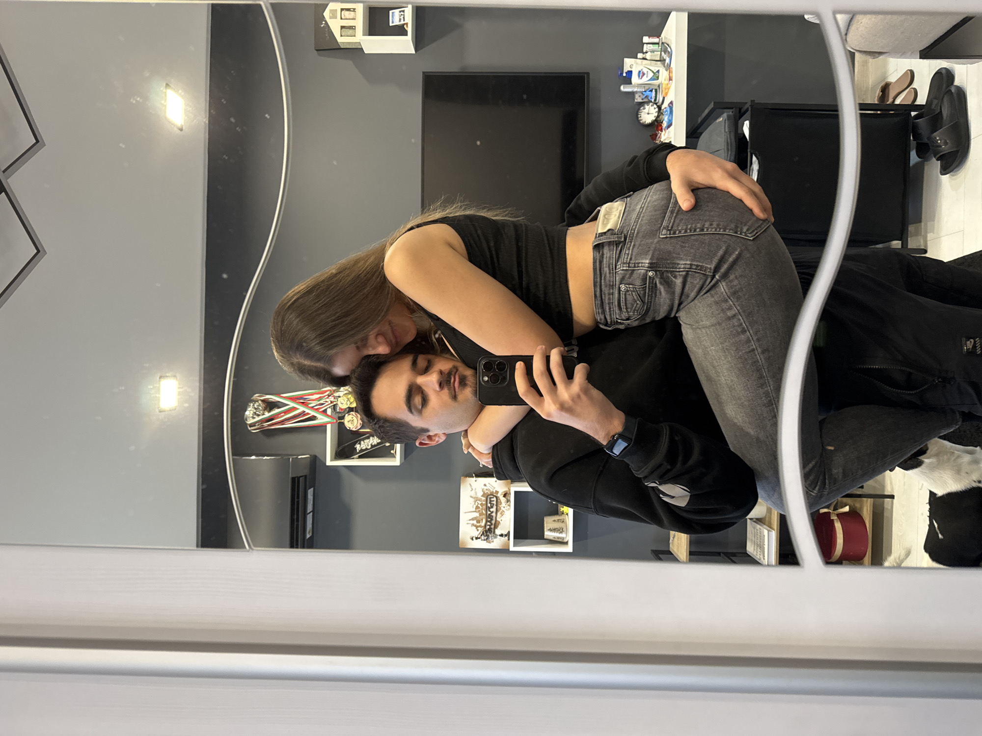 A man and a woman are posing for a selfie in a mirror. The woman is standing behind the man and has her arms wrapped around him. She has long brown hair and is wearing a black top and gray jeans. The man is wearing a black hooded sweatshirt and jeans, and he is holding a smartphone to take the picture. They are in a room with light gray walls, a black television, and shelves with various items. It is not possible to verify the location of the room in Sofia, Bulgaria based on the visual information provided.