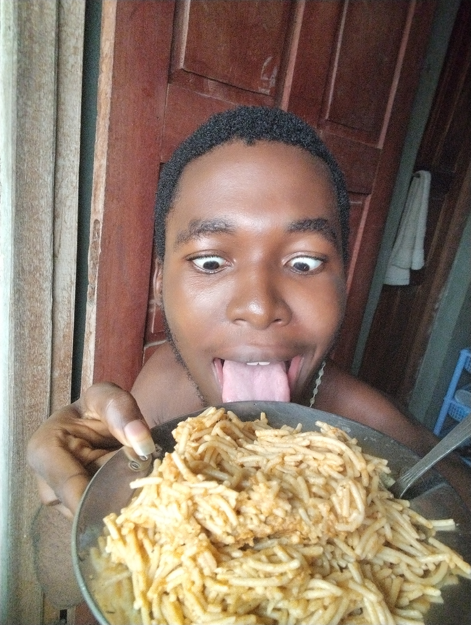 In Ihiagwa, Nigeria, a young person with dark skin and short, dark hair is looking into the camera with wide eyes. Their tongue is sticking out, and they are holding a metal plate of spaghetti with their left hand. The plate is positioned close to the camera, partially obscuring the lower half of their face. The background shows wooden doors, one a reddish-brown color and the other lighter wood with vertical slats. A white cloth is hanging on the wall in the background. The overall impression is of a candid or playful moment captured in what appears to be a home environment.