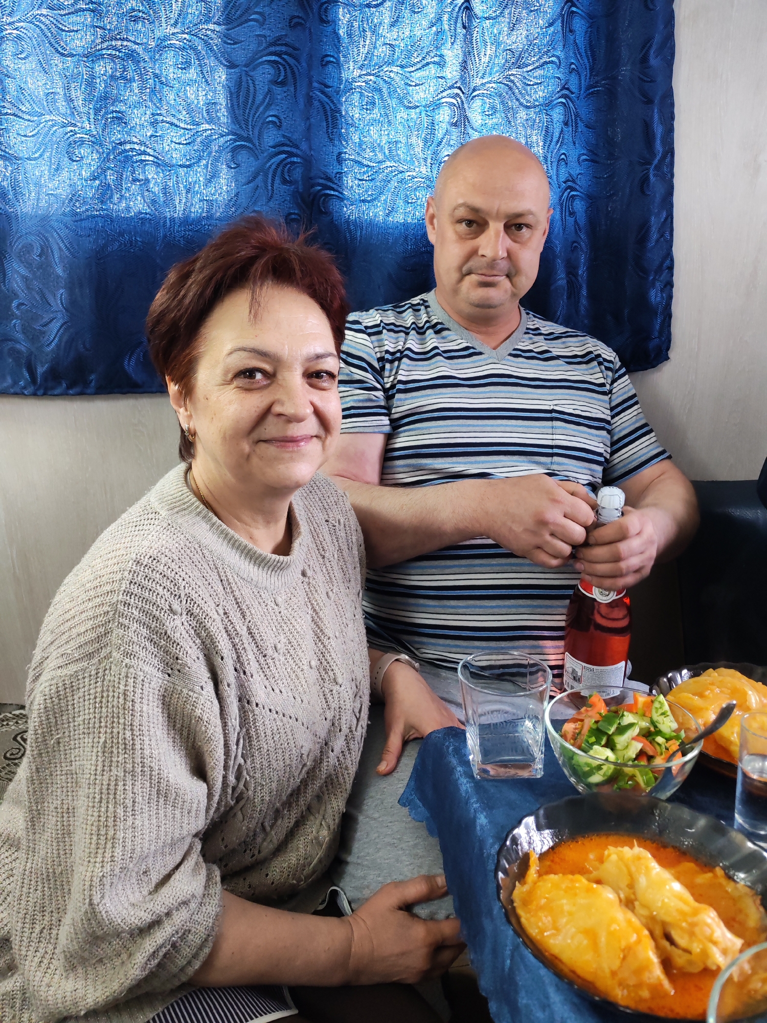 The image shows two individuals, a man and a woman, seated at a table in an indoor setting, likely a home in Shcheleiki, Russia. The woman, on the left, has short reddish-brown hair and wears a light-colored knitted sweater, smiling warmly at the camera. The man, on the right, is bald and wears a striped blue, white, and black t-shirt. He looks towards the camera with a neutral expression while his hands are busy opening a bottle containing a red liquid. The word "кВАСС" (Kvass) is faintly visible on the white label of the bottle, suggesting a traditional beverage.
The table in front of them is covered with a dark blue tablecloth and set with a clear glass, a bowl of fresh salad (chopped cucumbers and tomatoes), and two dark bowls holding what appear to be stuffed cabbage rolls in an orange sauce, indicating a meal is being shared. The background features a textured blue curtain, possibly covering a window, and a plain light-colored wall. The lighting suggests it is daytime. The scene conveys a casual and intimate gathering.