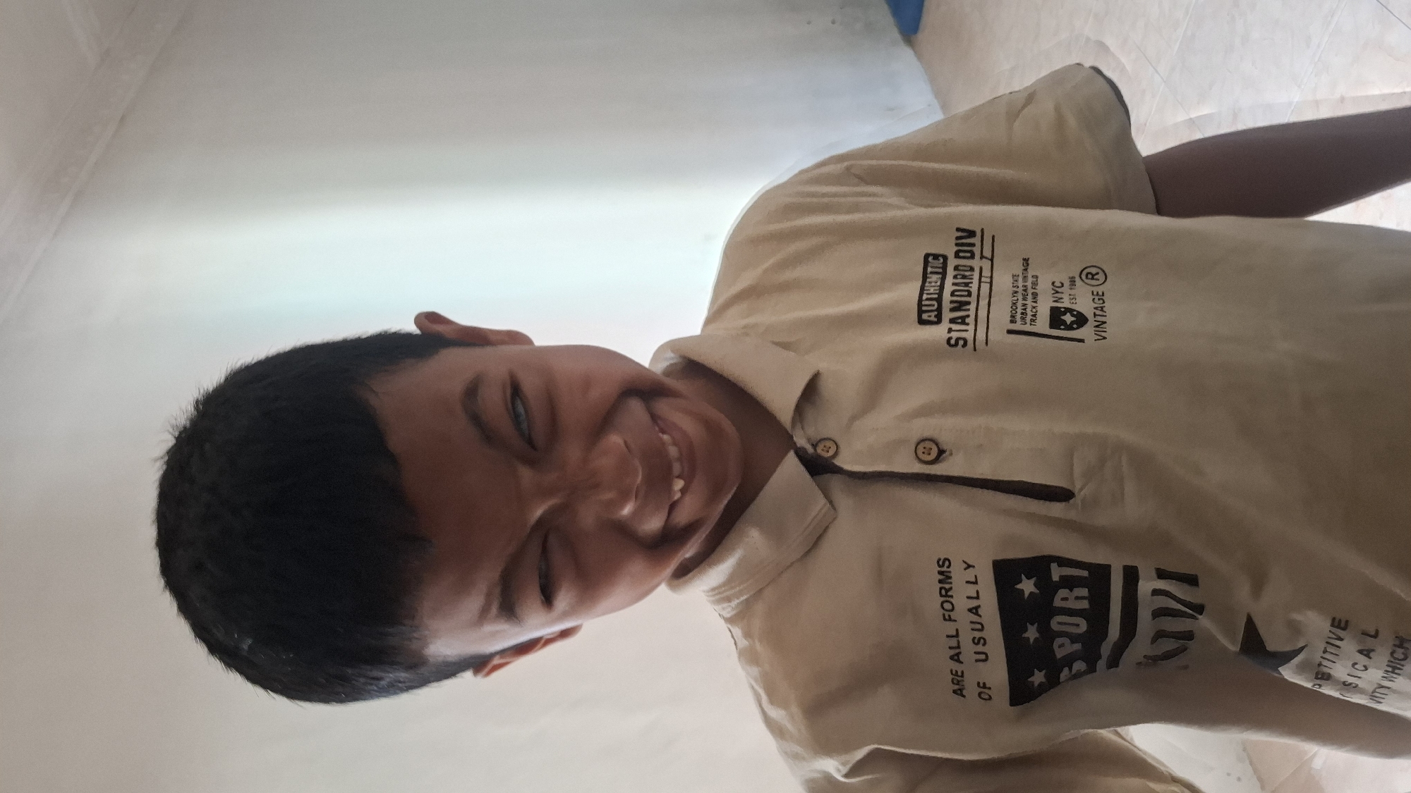 A young boy with dark hair and light-colored eyes makes a playful, slightly cross-eyed expression at the camera. He is wearing a light beige collared shirt with dark text and graphics. The text on the left side of his shirt reads "ARE ALL FORMS OF USUALLY SPORT". On the right side, it says "AUTHENTIC STANDARD DIV BROOKLYN STATE URBAN WEAR VINTAGE TRACK AND FIELD NYC EST. 1985 VINTAGE". He is positioned in an indoor setting, likely a home, with a plain, light-colored wall behind him and a tiled floor visible at the bottom of the frame. The lighting suggests it is daytime. The overall atmosphere is casual and informal, with the boy appearing to be interacting playfully with whoever is taking the photograph. This scene is located in the city of Cibinong Hilir, Indonesia.