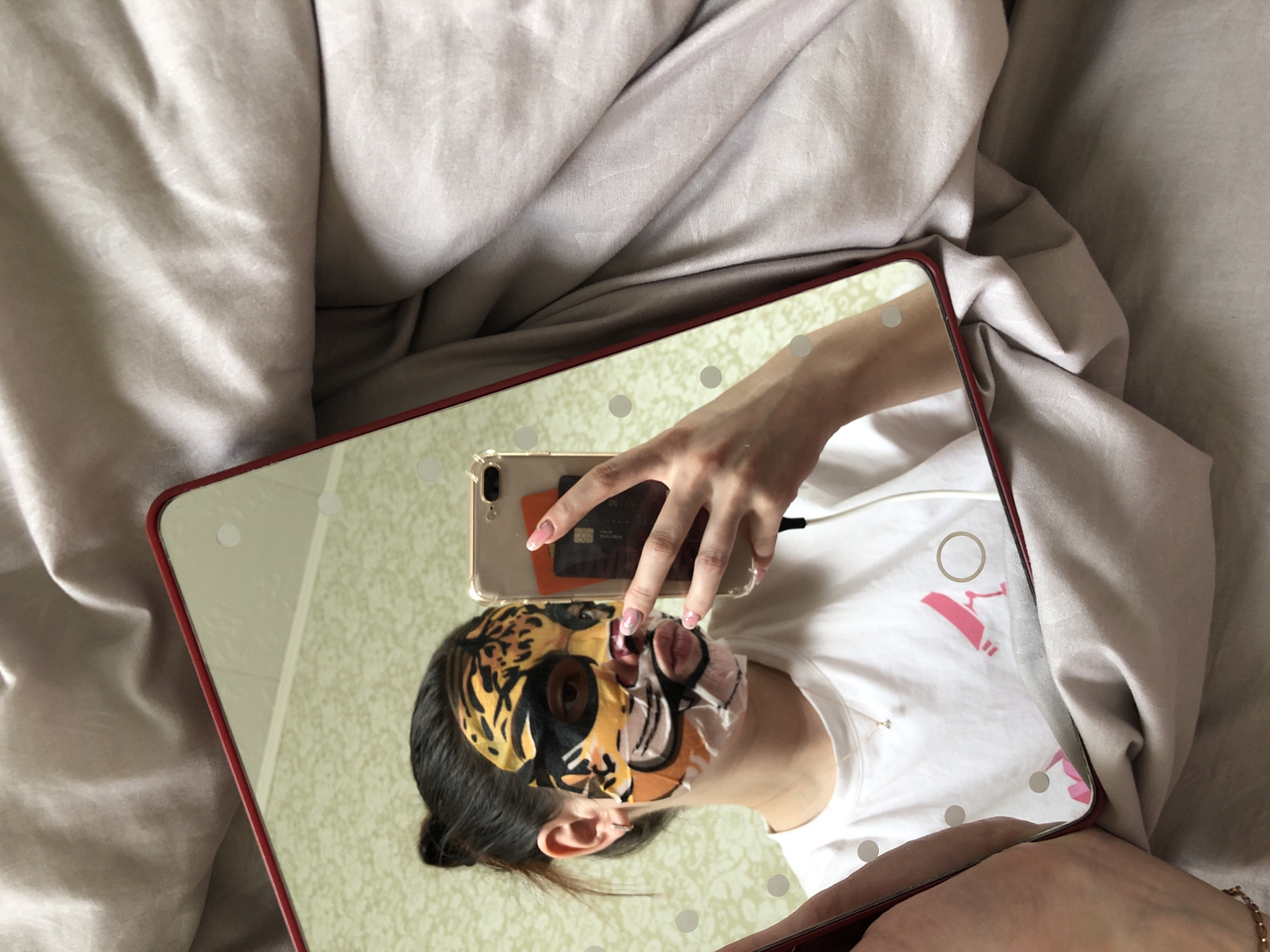An overhead photograph captures the mirror reflection of a person engaged in a self-care routine, likely in a bedroom setting in Noviy, Russia. The person, with dark hair and an ear piercing, is wearing a white t-shirt and a distinctive tiger-print sheet mask that covers most of their face. Their right hand holds a gold-colored smartphone, possibly an iPhone, encased in a clear cover. Several cards, including what appears to be a bank card with a black and gold design, are visible tucked behind the phone; faint text possibly includes "UNIONPAY". A black charging cable is plugged into the phone.
The mirror itself has a red frame and features several circular lights around its edges, suggesting it's a lighted vanity mirror. The person is reflected against a backdrop of light-colored, patterned wallpaper or a surface, while outside the mirror's frame, rumpled light beige or grey bedding is visible, indicating they are lying on a bed. A delicate gold necklace with a small pendant rests on the person's chest. The white t-shirt features a pink graphic, possibly a stylized letter "A" or an abstract design, along with a fainter white ring graphic above it. The scene conveys a private moment of relaxation and skincare.