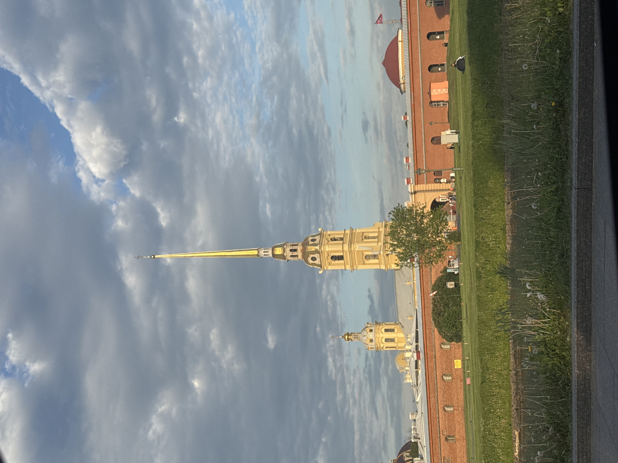The image, when correctly oriented, features the iconic golden spire of the Peter and Paul Cathedral in Saint Petersburg, Russia, dominating the center-left. The intricate gold spire, topped with an angel figure, rises prominently against a partially cloudy sky. Below and slightly to the right of the main spire, another smaller golden dome or tower, part of the same architectural complex, is visible.
To the right, a long, reddish-brown brick wall, characteristic of the Peter and Paul Fortress, stretches across the midground. Behind this wall, a red-domed building, likely within the fortress grounds, stands out. A flagpole with a flag, possibly the St. Andrew's flag, is visible above this building. Several arched windows and lampposts punctuate the brick wall.
The foreground is dominated by a wide, green grassy area dotted with dandelions and other wild plants. A single individual is seen sitting on the grass near the brick wall, engaged in leisure. The sky features a mix of white and grey clouds, with patches of clear blue visible through breaks in the cloud cover. The overall lighting suggests it is daytime, possibly late afternoon, with bright, diffused light illuminating the scene. No discernible text is visible on any signs or structures. The image captures a serene moment at a historic landmark, highlighting its architectural beauty and natural surroundings.