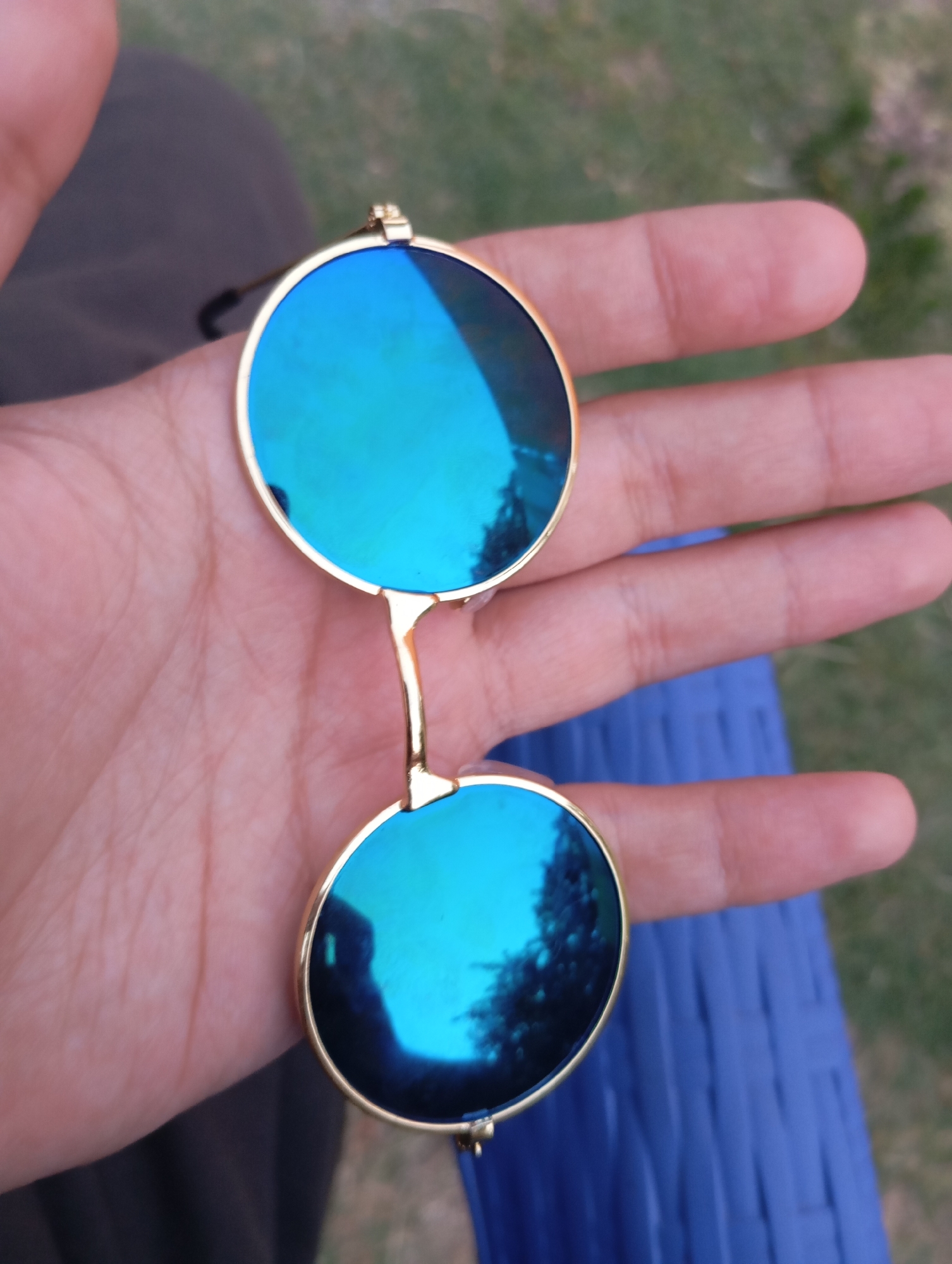 A pair of round, gold-rimmed sunglasses with bright blue mirrored lenses are being held in a person's hand. The sunglasses are held with the lenses facing upwards, reflecting a blurry blue and green environment. The hand is slightly cupped, and the sunglasses are positioned centrally within the palm. In the background, out of focus, is green grass and a textured blue surface, possibly a chair or fabric.