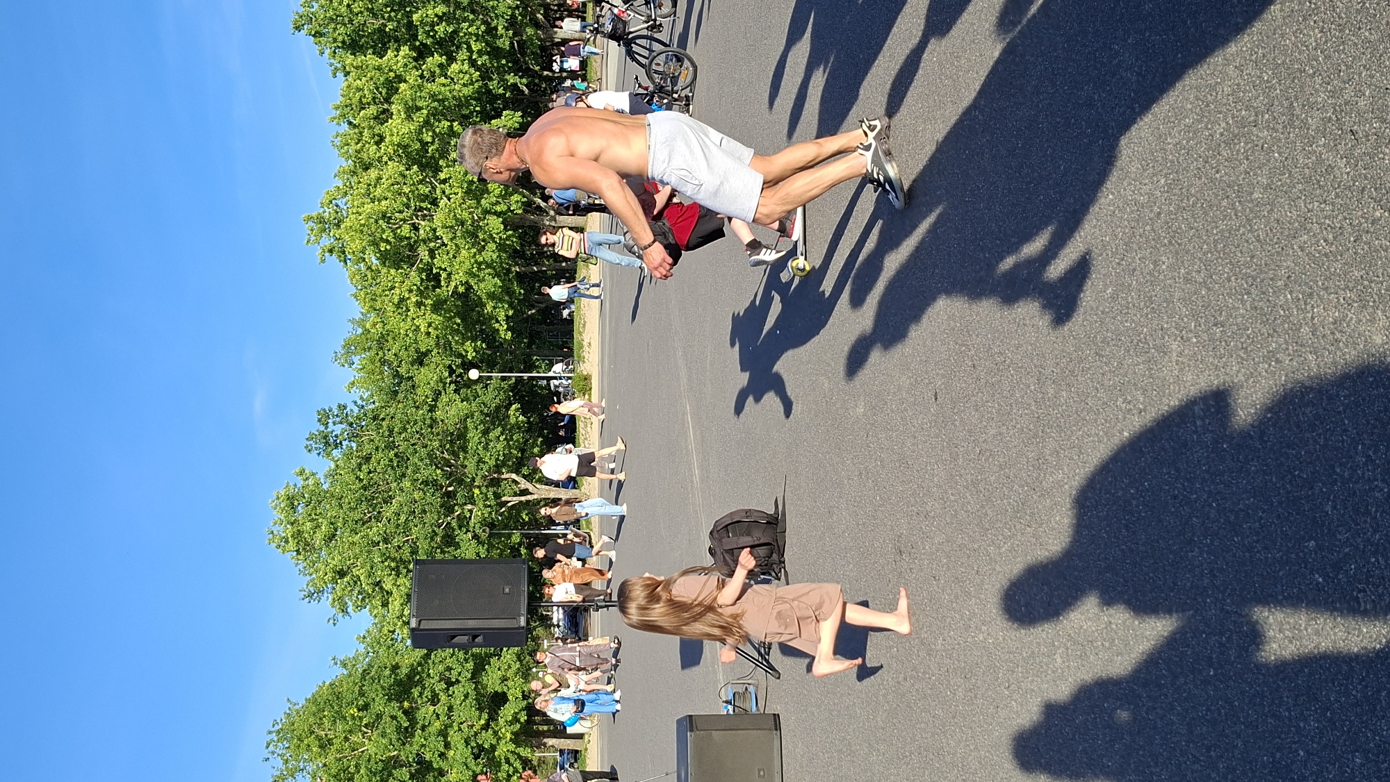 An outdoor scene in Tallinn, Estonia, features a clear blue sky and lush green trees lining a paved area. In the foreground, a young barefoot girl in a brown dress runs towards the left of the frame. To her right, a shirtless man wearing light-colored shorts and sneakers is in motion, appearing to interact with a person on a skateboard. A large black speaker is positioned on the left side of the image, with another speaker and a black backpack near the running girl. Several other people are visible in the background, some walking and others engaged in activities, including a person on a bicycle. Shadows of people are cast on the pavement.