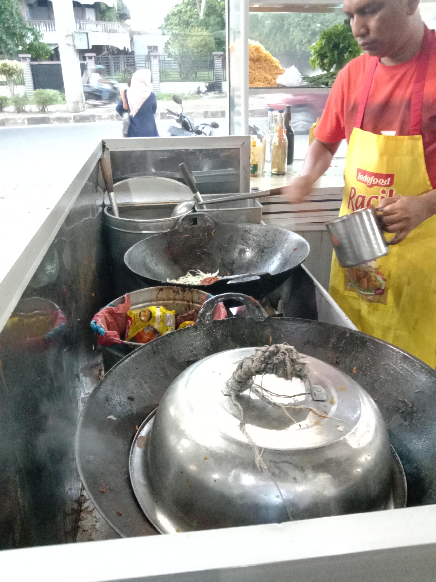 A street food vendor in Lhokseumawe, Indonesia, wearing a red shirt and a yellow Indofood apron, is preparing food. He holds a metal cup in his right hand, near a large, dark metal wok. In front of him is another wok, covered with a metal lid secured by rope. Behind the vendor, bottles and stacked noodles are visible. In the background is a street with a passing pedestrian in a headscarf and a parked motorcycle, and across the street are buildings and trees.