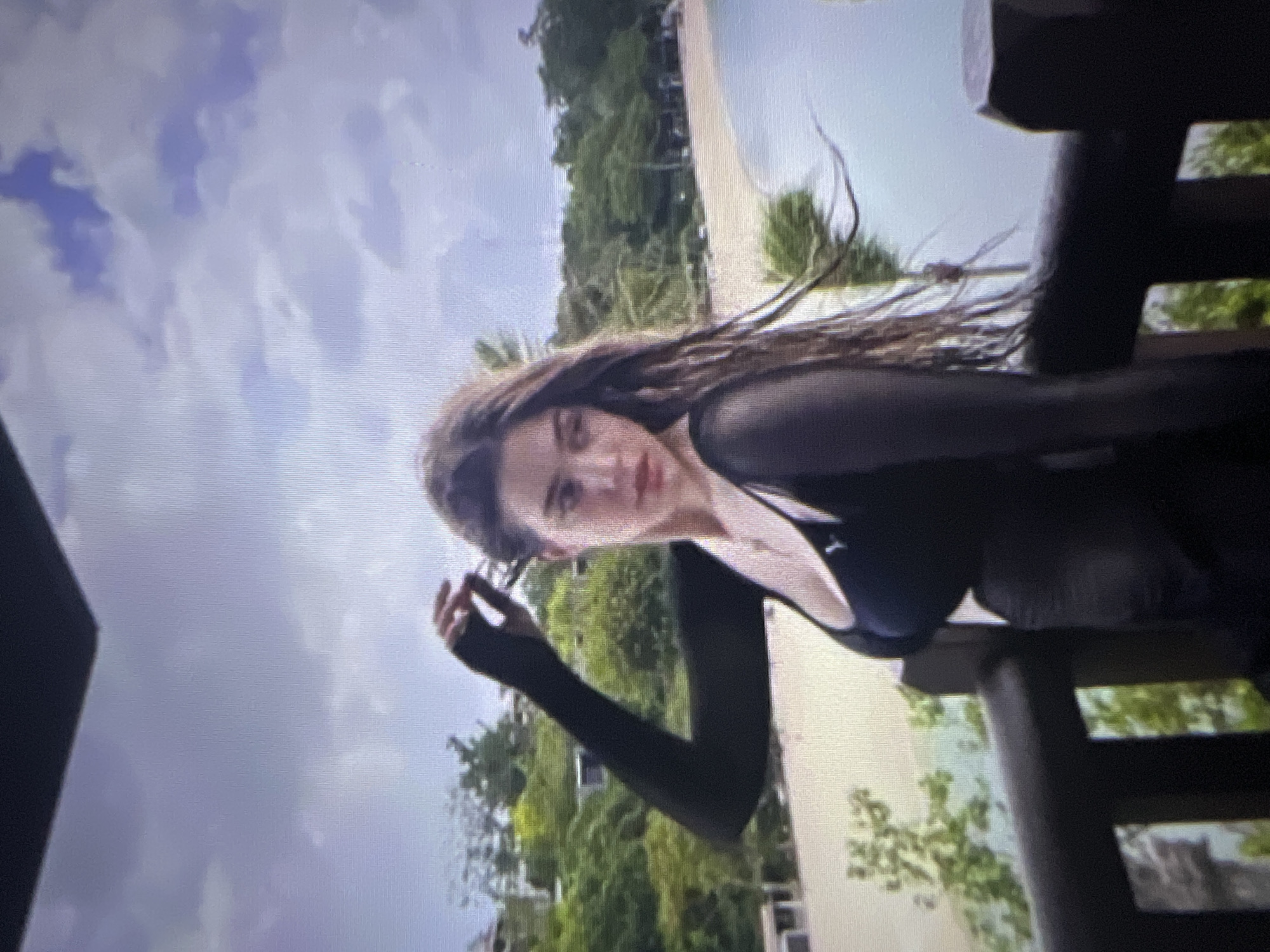 A woman with long, dark, wavy hair stands on a wooden balcony, looking to her right. She is wearing a black top with long sleeves and a neckline that reveals her chest. She has a necklace on. Behind her, there is a beach with light-colored sand and blue water, bordered by lush green trees. The sky is cloudy and overcast. The image quality is low, with visible pixels and a grainy texture. The location is not specified.