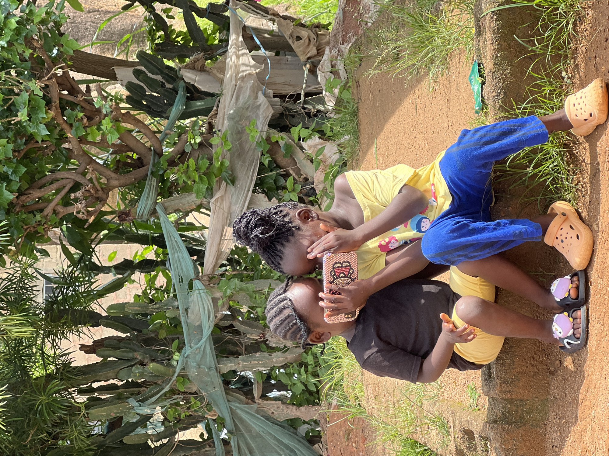 Two young children are sitting on a low, rough-hewn stone wall outdoors. The child on the left, wearing a black t-shirt and yellow shorts, is holding a pink, jeweled smartphone in both hands, looking down at it. Their hair is styled in neat braids. The child on the right, wearing a yellow off-the-shoulder top and blue pants, is partially covering their face with their hands, seemingly looking at the phone as well or reacting to its content. Their hair is in braided pigtails. Both children are wearing sandals. The ground in front of them is reddish-brown dirt. Behind them, a dense, overgrown area of trees and large cacti are visible. Green and white plastic sheeting is draped over some of the plants. The overall setting suggests an outdoor, possibly informal, residential area in Jos, Nigeria. The lighting indicates it is daytime.