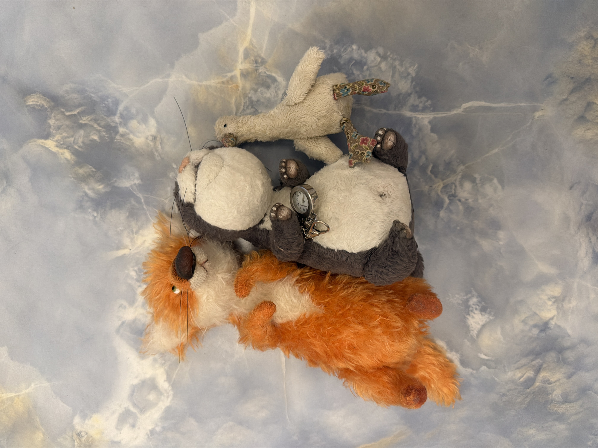 Three stuffed animals are arranged in the center of the frame against a textured blue and white background. On the left is an orange fox with a white chest and inner ears, with its front paws resting on its belly. In the middle is a grey and white cat-like stuffed animal with a pocket watch attached to a chain around its neck. On the right is a small, light-colored stuffed goose with colorful floral pattern patches on its legs and feet. The background appears to be a marble-like surface with blue and white veining.