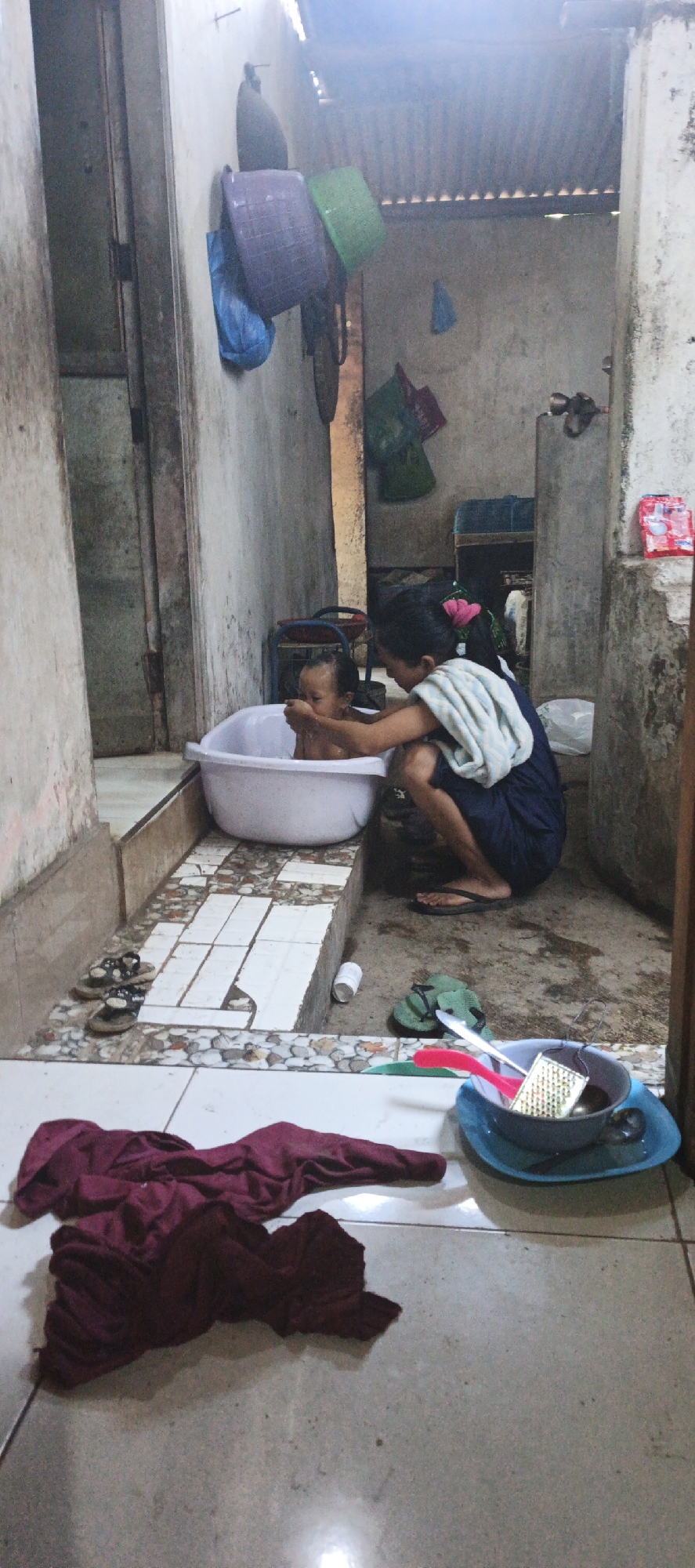 In Kasiyan, Indonesia, a young person is bathing a toddler in a white plastic tub, positioned on a tiled platform. The caregiver, wearing a dark blue dress and a striped towel draped over their shoulders, crouches beside the tub, tending to the child. A pink flower is in the caregiver's hair. The environment appears to be an outdoor or semi-outdoor bathing area with a corrugated metal roof visible. To the left, a doorway with a wooden frame leads into another space, and a wall is adorned with several woven baskets. On the floor in the foreground, a pile of maroon fabric is spread out, and a blue plastic basin holds a grater, a spoon, and a red utensil. Green flip-flops and another pair of sandals are also present on the tiled area. The scene suggests a domestic, utilitarian setting.