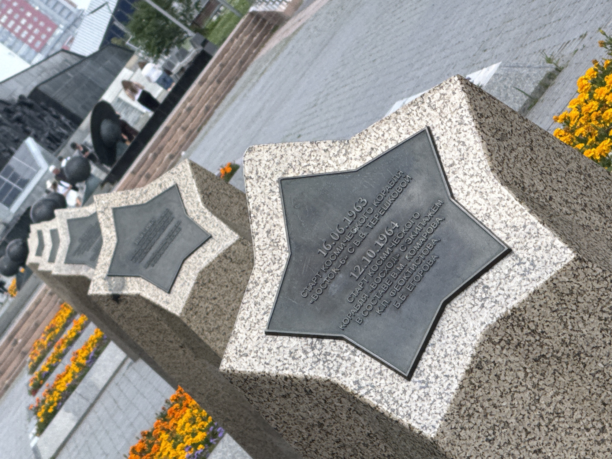 The image depicts a monument with star-shaped reliefs, likely commemorating space exploration. The monument is made of granite and features inscribed plaques in Russian. The first plaque, dated "16.06.1963," refers to the start of the "Vostok-6" space mission with V.V. Tereshkova. The second plaque, dated "12.10.1964," mentions the start of the "Voskhod" mission with crew members V.M. Komarov, K.P. Feoktistov, and B.B. Yegorov. The monument is situated in an outdoor setting with pathways, flower beds containing orange and purple flowers, and stairs in the background. Other people are visible in the background, some standing near the monument and others further away, near buildings that appear to be part of a city environment. This monument is located in Moscow, Russia, and is part of the Moscow State Memorial Museum of Cosmonautics or a similar space-themed attraction, possibly near VDNKh.