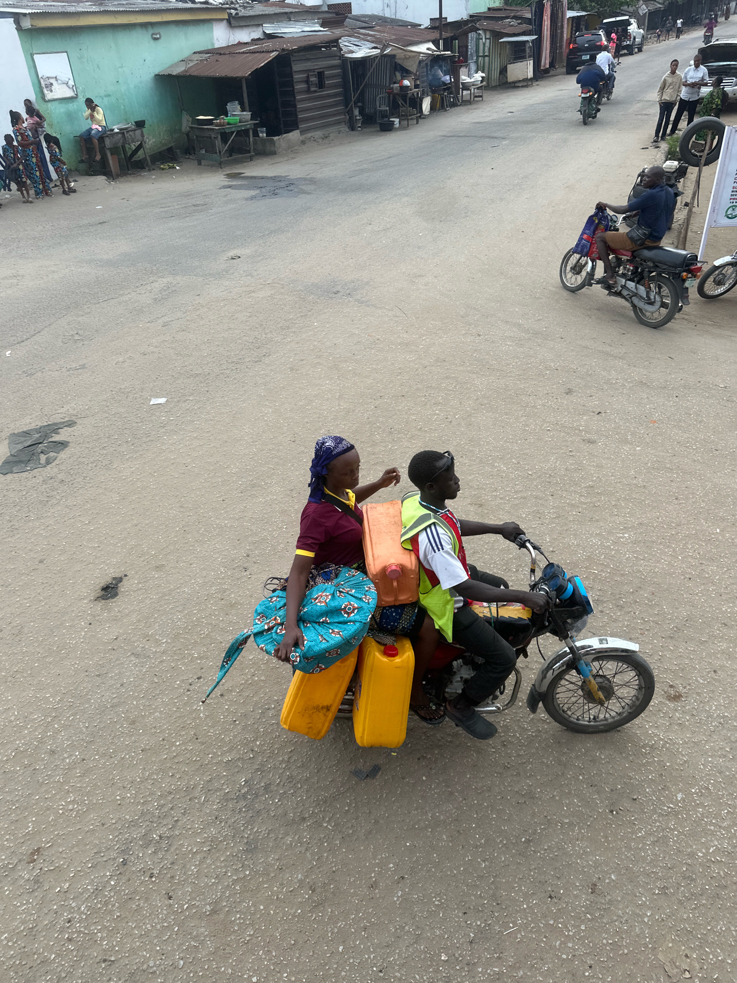 In Ajara, Nigeria, a man and a woman are riding a motorcycle on a paved road. The man is driving, wearing a yellow vest over a white and red striped shirt and black pants. The woman is the passenger, seated behind him, wearing a maroon shirt, a blue and white headscarf, and a patterned blue and white wrap skirt. The motorcycle is loaded with three large yellow containers and a blue and white patterned bag. The surrounding environment includes buildings with corrugated metal roofs, some appearing to be shops or homes, and other vehicles, including another motorcycle in the distance. The road surface is rough asphalt. Several other individuals are visible in the background, standing and walking near buildings.