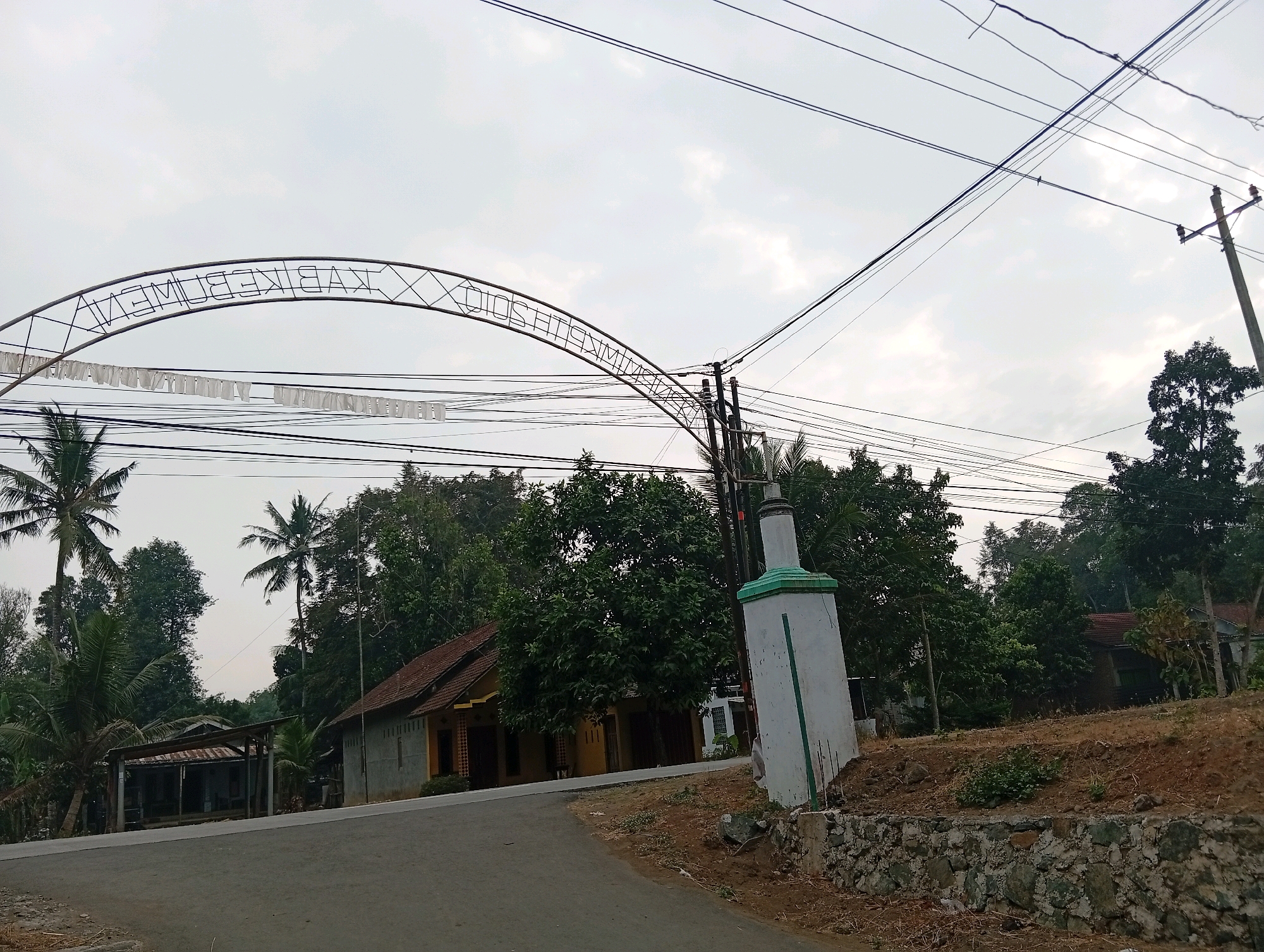 The image shows an arched metal structure over a road in Plempukankembaran, Indonesia. The structure appears to be an entrance gate with intricate metalwork and a sign that reads "KEMBARAN". Below the arch, a string of white fabric banners is hung. Several utility wires crisscross the sky above the arch and road. To the left of the road, there is a small, single-story building with a brown tiled roof and a shaded porch area. Further back, lush green trees and palm trees are visible. On the right side of the road, a white rectangular structure with a green trim stands near the road, possibly a monument or marker. Behind this structure, a dry, brown, and rocky embankment slopes upwards, dotted with some vegetation. Another house with a brown tiled roof is visible in the background on the right, partially obscured by trees. The sky is overcast with a pale, diffused light, suggesting it is either early morning or late afternoon. The scene depicts a rural or semi-rural environment.
