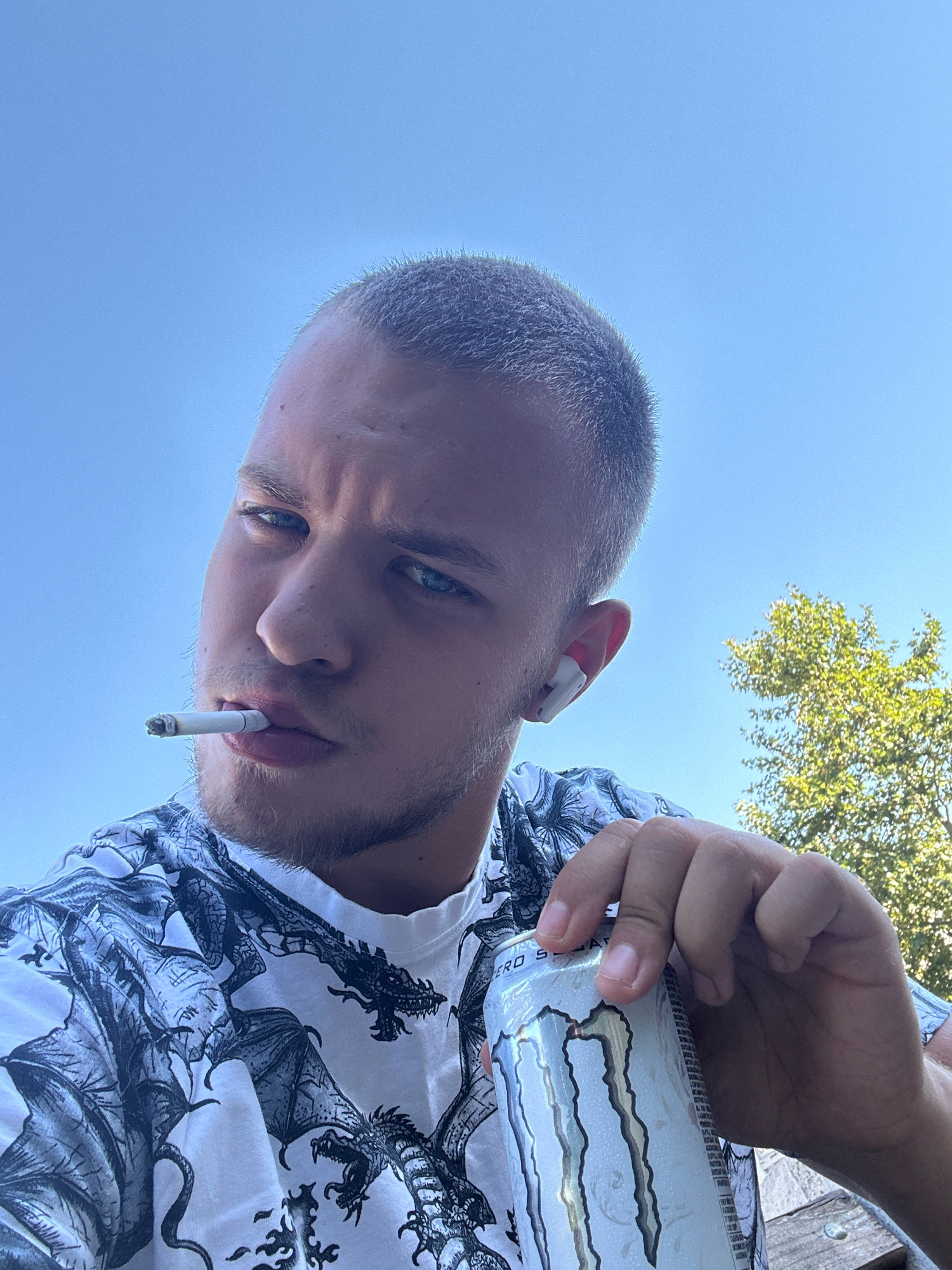 A close-up, low-angle selfie shows a young man with short, buzzed blond hair and piercing blue eyes, looking directly at the camera. He is holding a lit cigarette in his mouth, with smoke slightly obscuring his lips. He is wearing a white t-shirt with a black dragon print. In his right hand, he is holding a white can of Monster Energy drink. White wireless earbuds are visible in his right ear. The background is a clear blue sky with the upper branches of a lush green tree visible on the right side. The image appears to be taken outdoors on a sunny day. There is no identifiable location in the city of Sliven, Bulgaria present in the image.