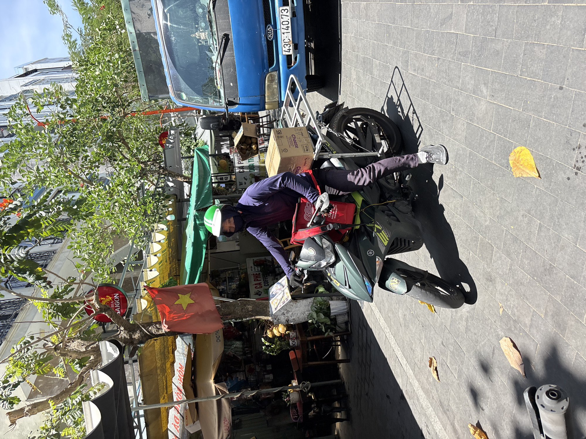 A person wearing a purple long-sleeved shirt, dark pants, and a green helmet rides a dark green motorcycle on a paved street in Đà Nẵng, Vietnam. The motorcycle has a silver rack on the back holding a cardboard box. The rider is wearing a mask and gloves. A red bag is attached to the back of the motorcycle. In the background, there are several market stalls with awnings, a blue truck, and buildings. A Vietnamese flag is visible hanging from a tree. The ground is paved with grey stones, and fallen leaves are scattered on the ground. The scene appears to be a busy street in a city.
