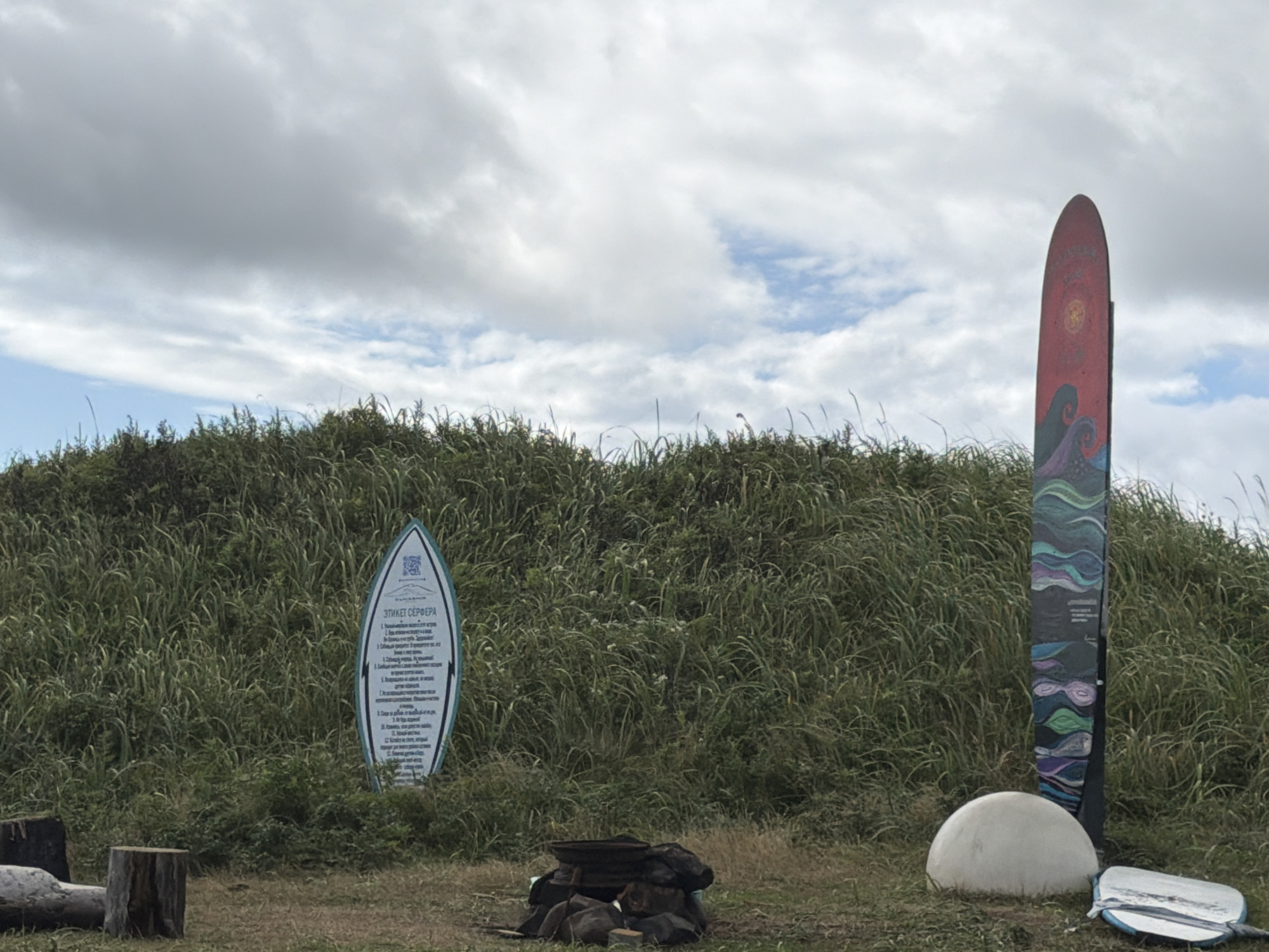 An outdoor scene on a cloudy day features a grassy hill in the background with a tall, thin, surfboard-shaped sign on the right side of the image, adorned with colorful wave-like patterns and illustrations. To the left of this sign, further back on the hill, is another surfboard-shaped sign with text in Russian, appearing to be a set of rules or guidelines. In the foreground, there is a round, white, dome-shaped object, and a white surfboard lying on the ground. Two wooden stumps and a log are also visible in the lower-left portion of the frame. The environment suggests a coastal or beachside location, likely in Yuzhno-Kurilsk, Russia, given the presence of Cyrillic script on one of the signs. No people are present in the image.