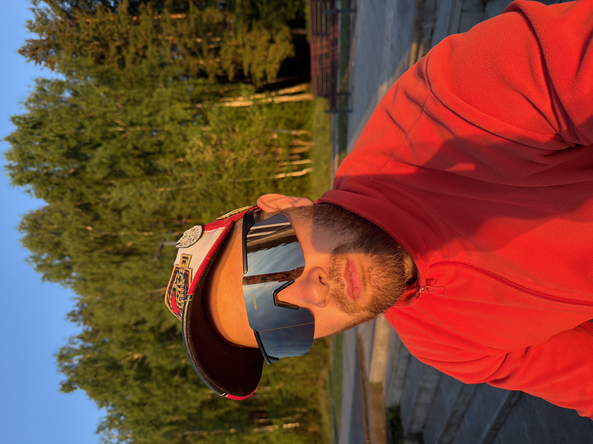A man wearing a red fleece jacket and a baseball cap with multiple patches and a silver emblem, and dark tinted sunglasses, is photographed outdoors in Chaikovsky, Russia. He has a beard and is looking directly at the camera. The background shows a line of trees, a park bench, and stairs, suggesting a park or recreational area. The lighting indicates it is either early morning or late afternoon, with the sun casting a warm glow on the scene.