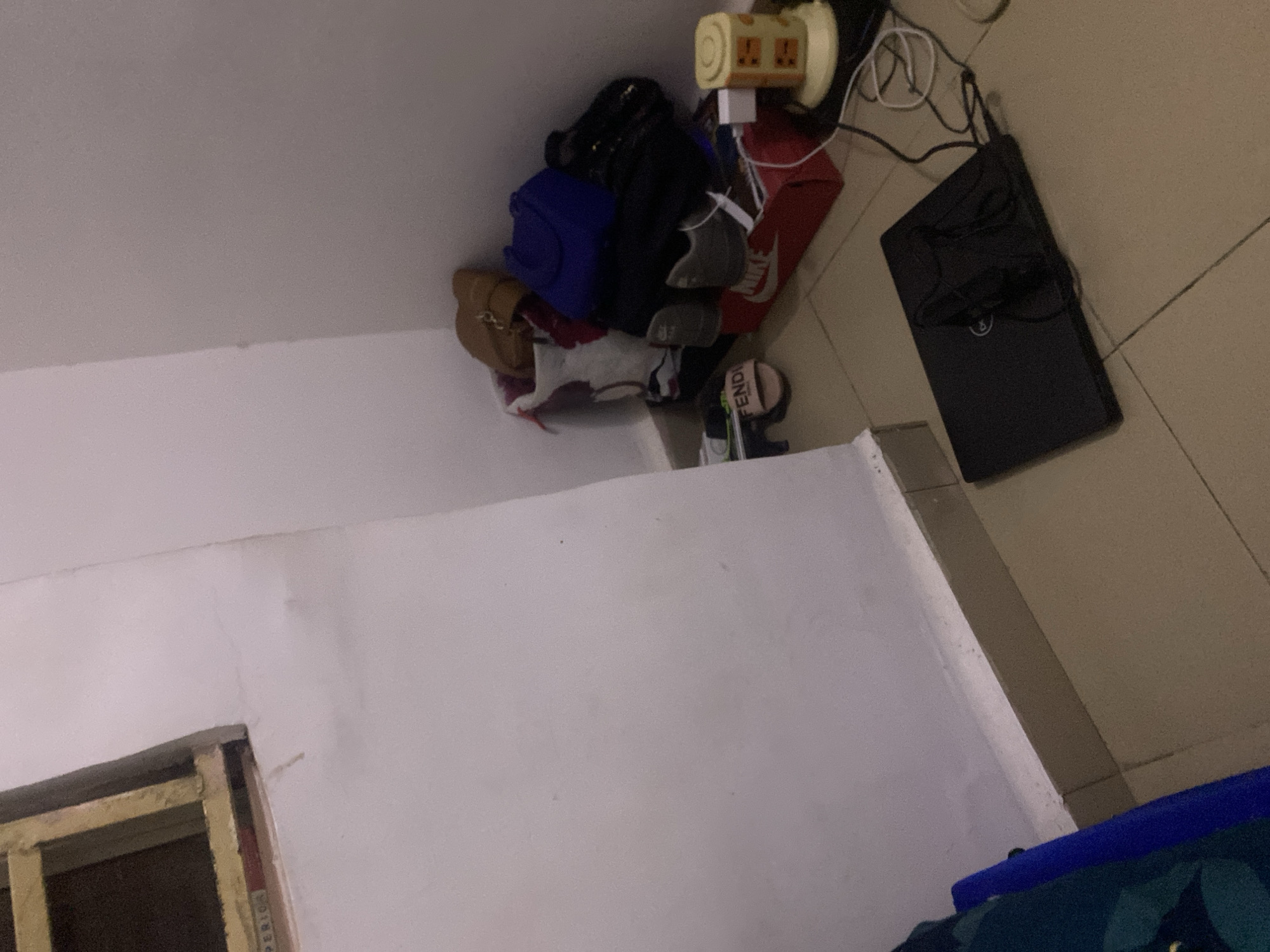 The image depicts an indoor corner of a room with tiled flooring. In the corner, a collection of items is piled up, including bags, shoes, and a red Nike shoebox. A pink Fendi sandal is visible near the bottom left of the pile. A black laptop is open on the floor in the foreground, with its charging cable connected. To the right of the pile of items, a small yellow and white electrical appliance, possibly a charger or adapter, is plugged into a power outlet. The walls are white and appear slightly textured, with some faint marks. There is a window frame with wooden slats visible in the upper left portion of the image. The overall impression is of a cluttered domestic space. There is no specific information in the image to indicate the location as Howayi, Nigeria.