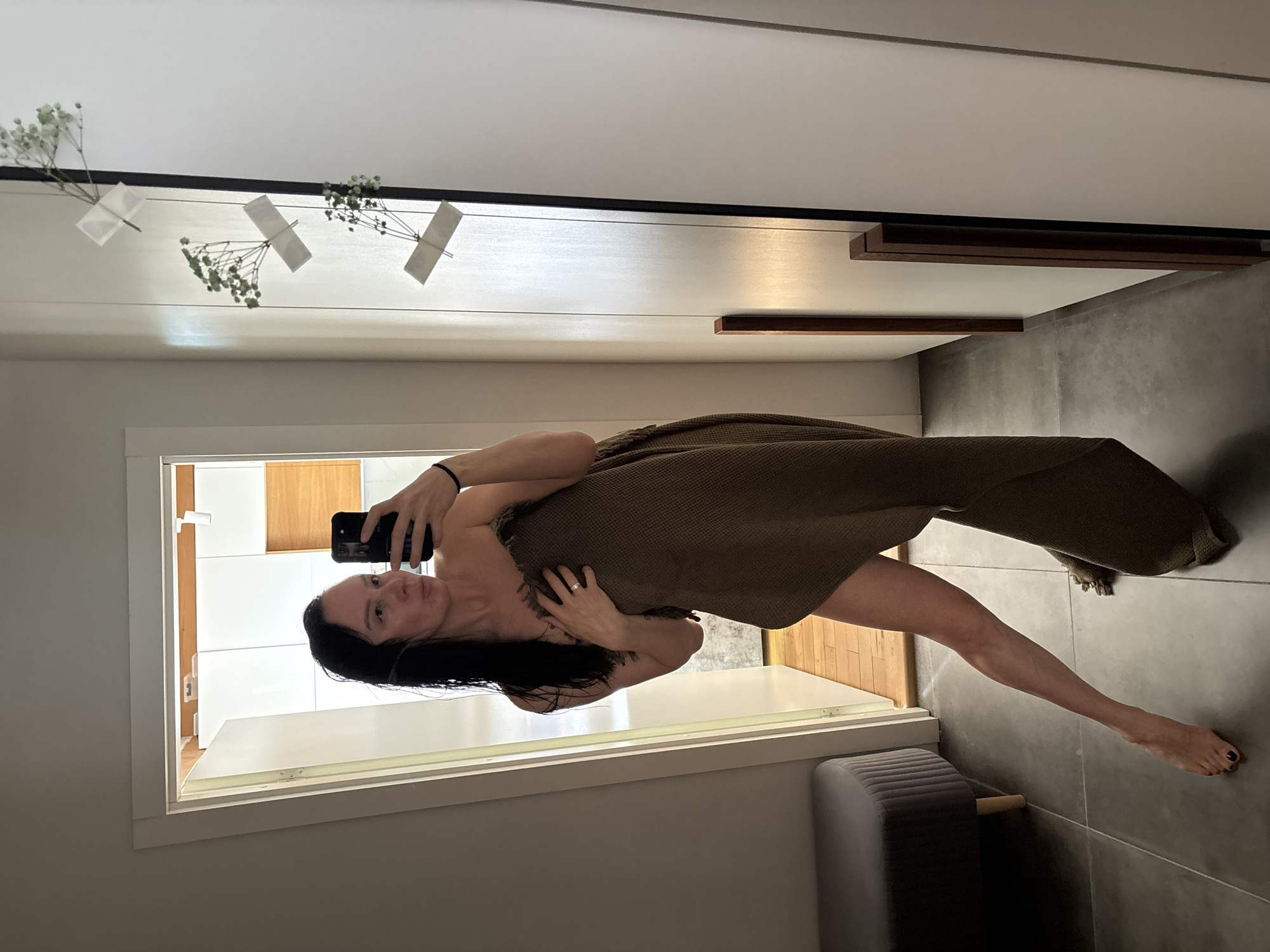 A woman with long, dark hair is taking a selfie in what appears to be a bathroom or dressing area. She is partially covered by a brown woven wrap or blanket. She is holding a black smartphone in her left hand, positioned to capture the image. The bathroom has a modern design with white and wood-paneled walls, and gray tile flooring. Small bouquets of baby’s breath are taped to the wall near the ceiling. The image suggests a private setting, possibly a home. There is no indication of a location.